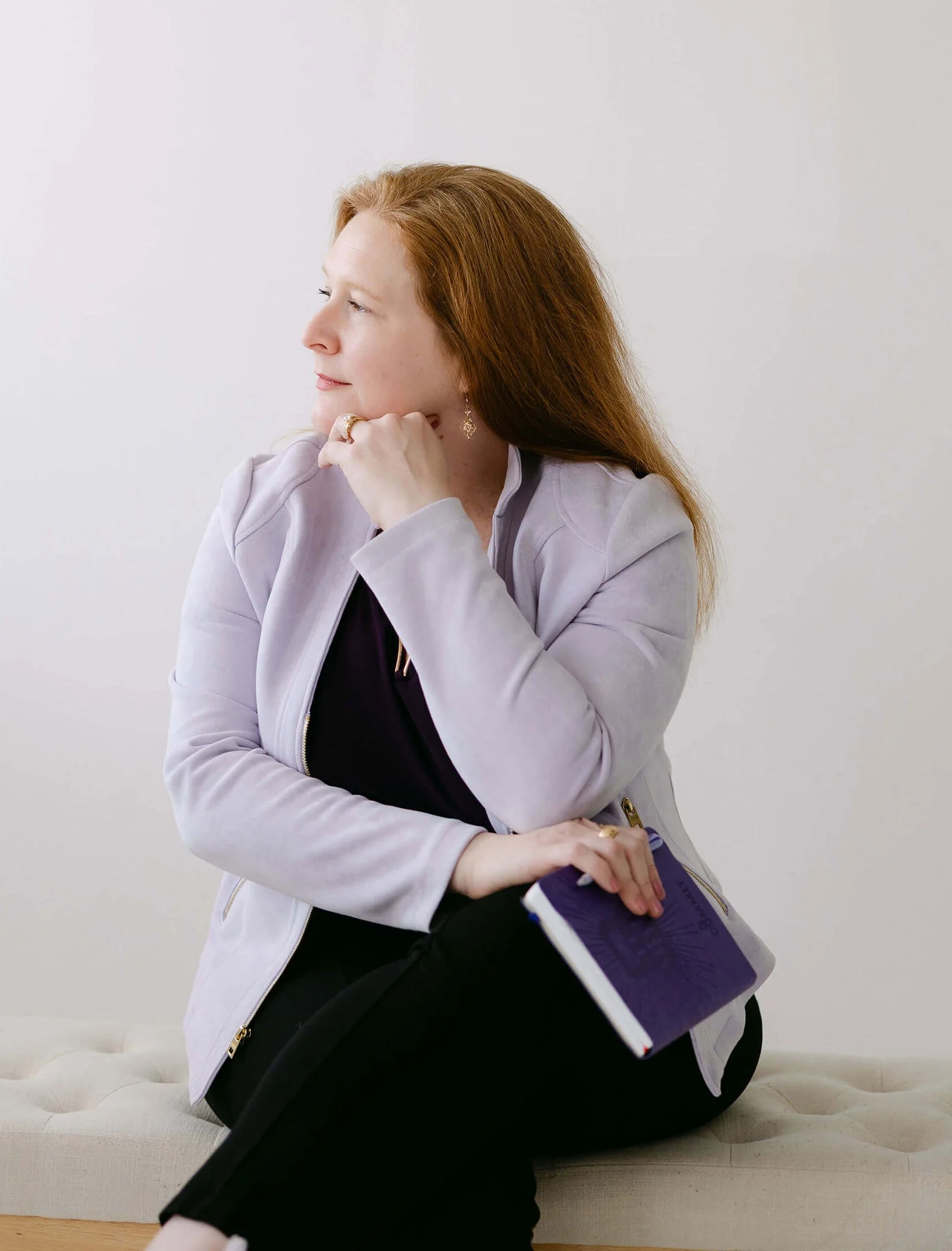 A woman with red hair, wearing a light gray jacket and jewelry, sitting on a beige sofa, holding a purple folder, and looking to her left against a plain white wall.