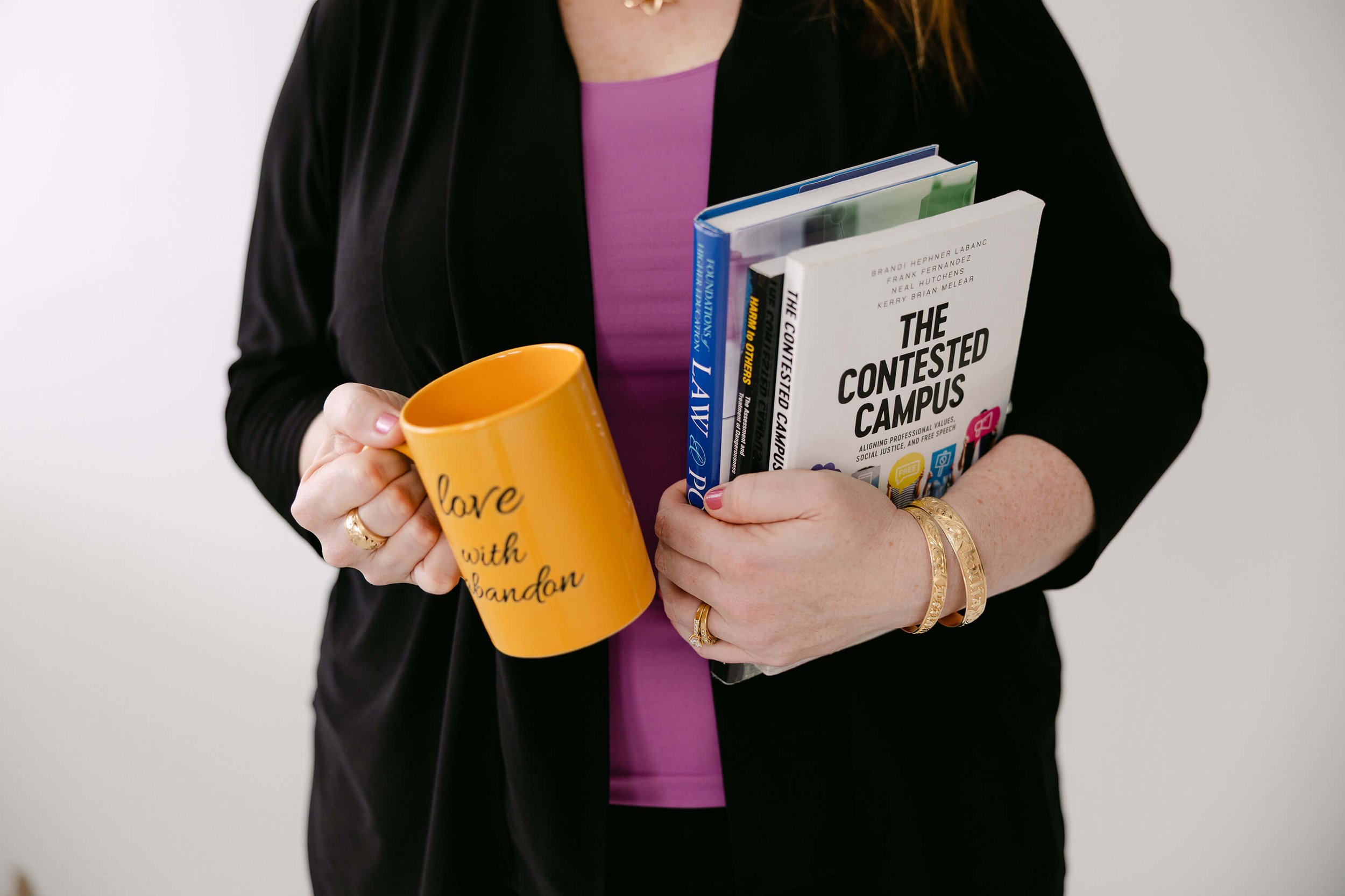 A woman wearing a black jacket and a purple top holding a yellow mug and several books. The mug has the phrase 'love with abandon' written on it. The books include titles like 'The Contested Campus' and others.