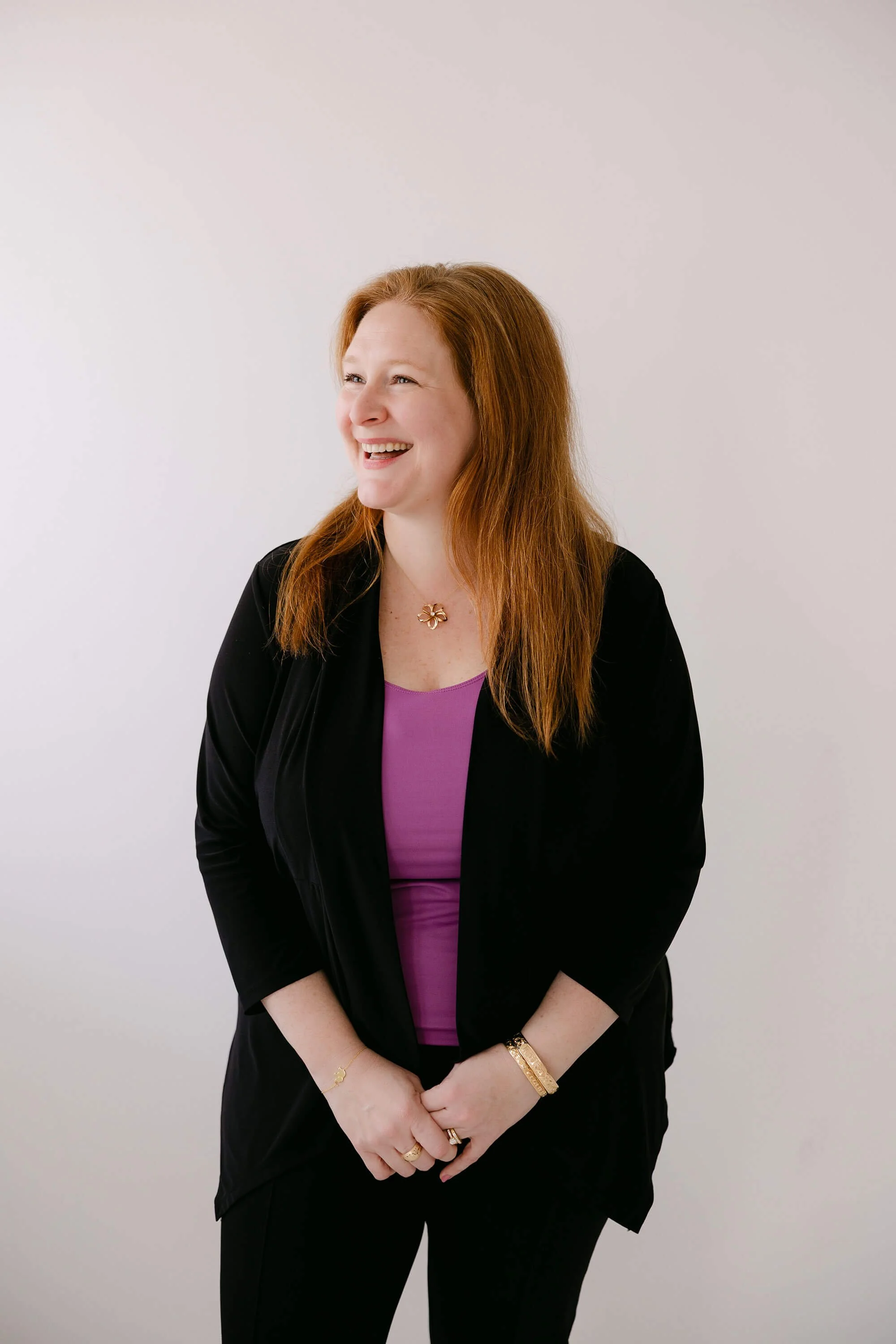 A woman with long red hair smiling and looking to her right, wearing a purple top, black blazer, and gold jewelry against a plain white background.