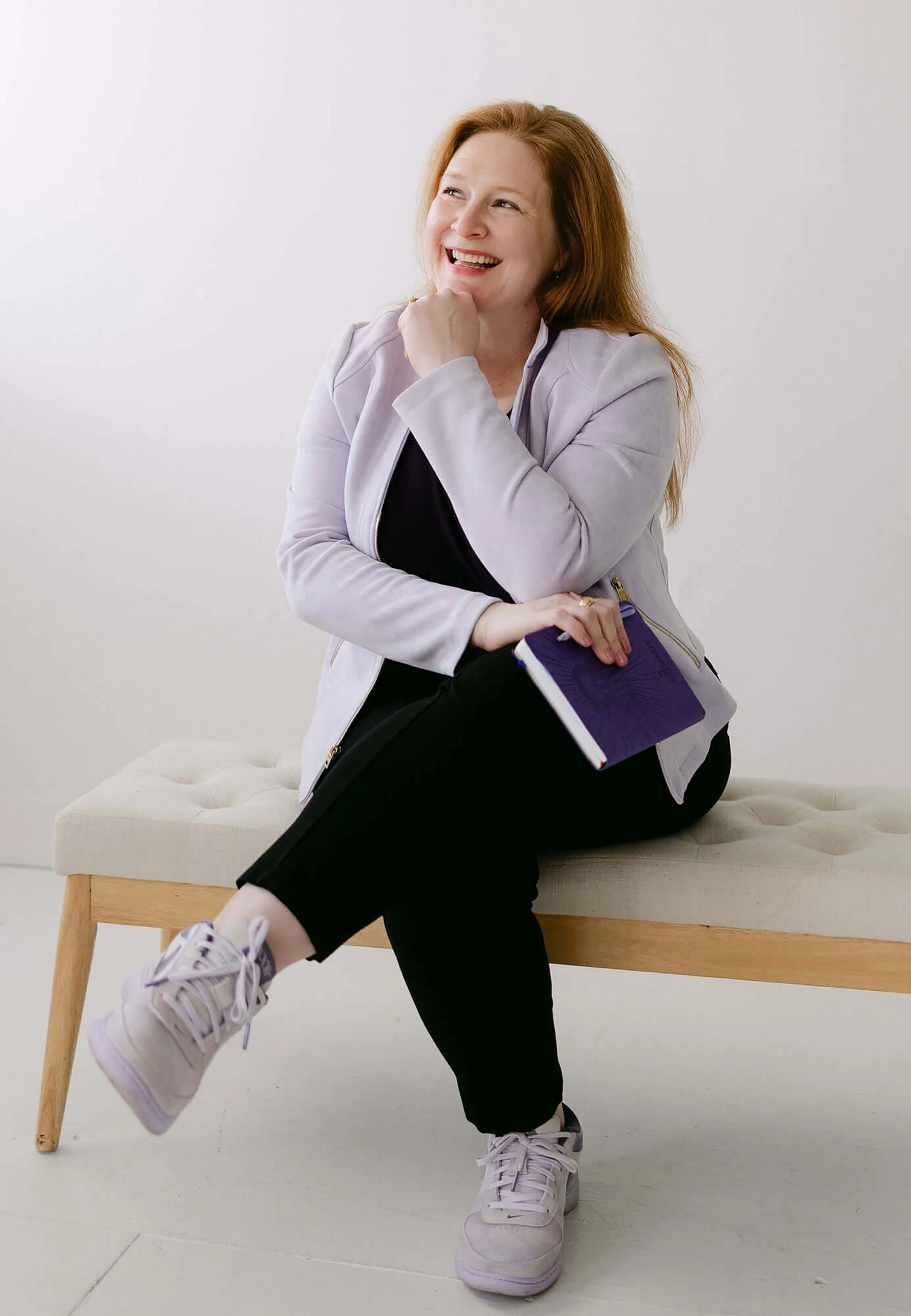 A woman with red hair, wearing a light gray jacket and black pants, sitting on a beige cushioned bench with wooden legs, holding a purple notebook, smiling and looking to her right.