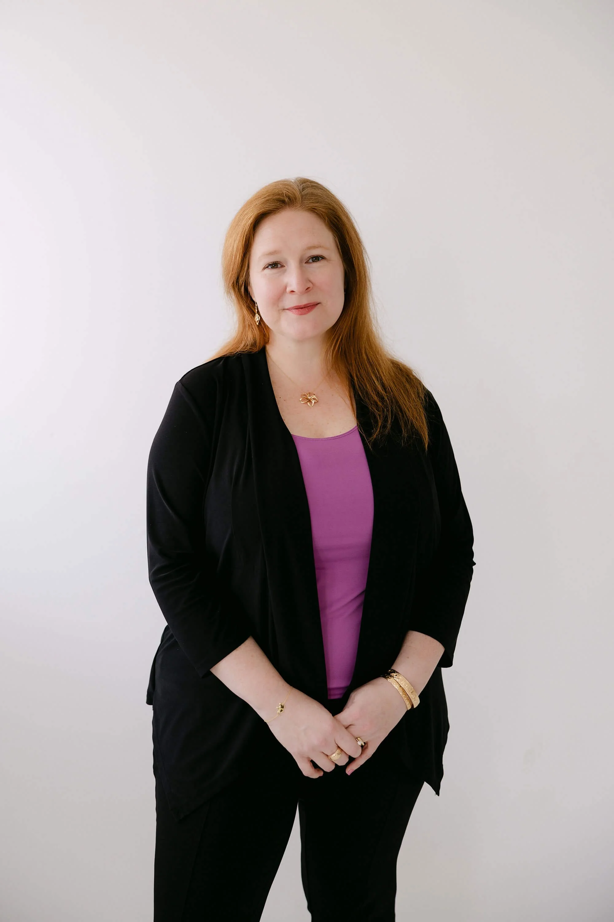 A woman with red hair wearing a purple top, black blazer, and black pants, standing against a white wall, smiling slightly.