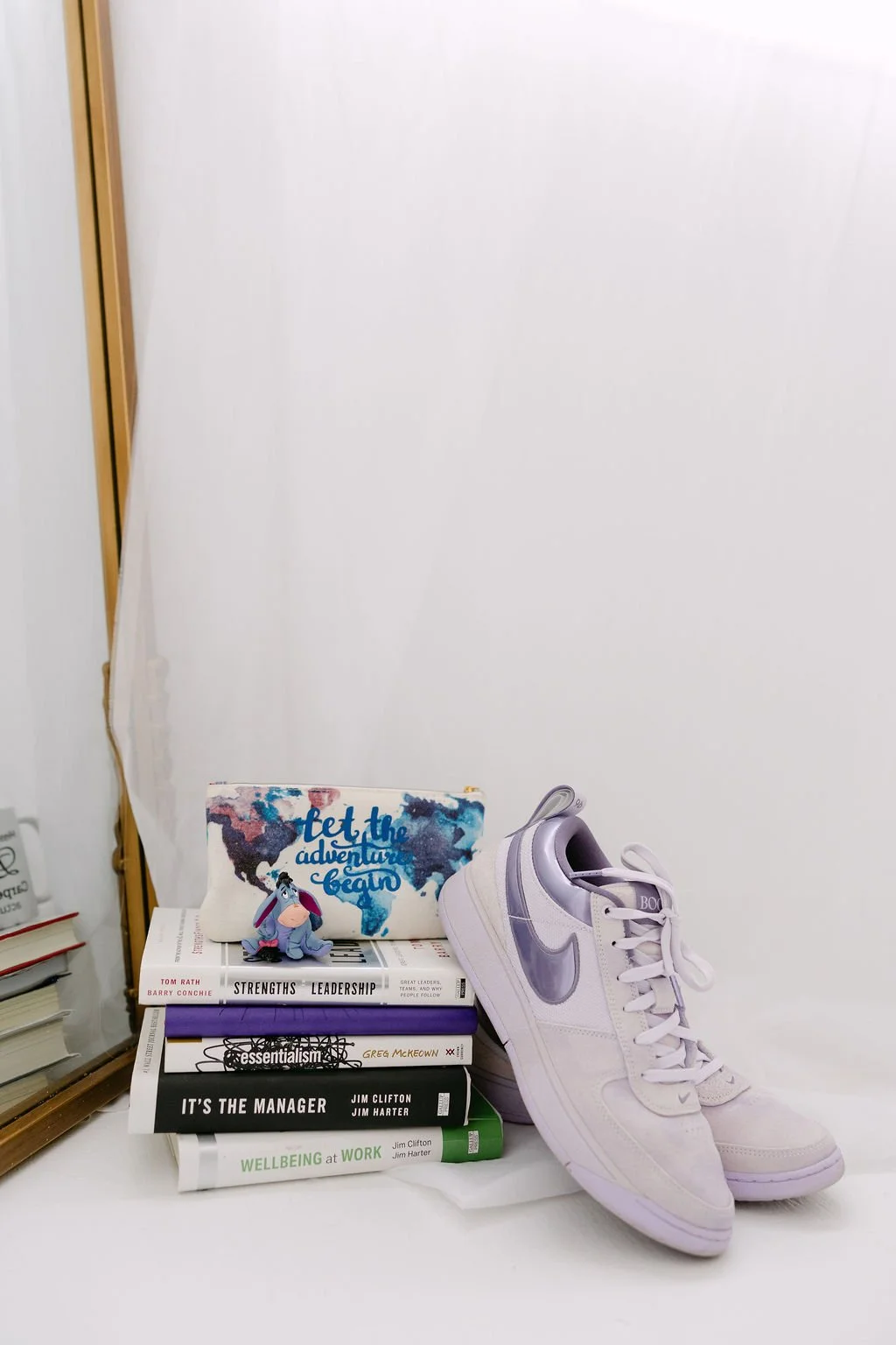 Stack of five books with a plush Eeyore toy on top, a white pencil case with a world map design and the phrase 'Let the adventure begin', and a pair of white Nike sneakers with silver swoosh logos placed on a white surface against a white background.