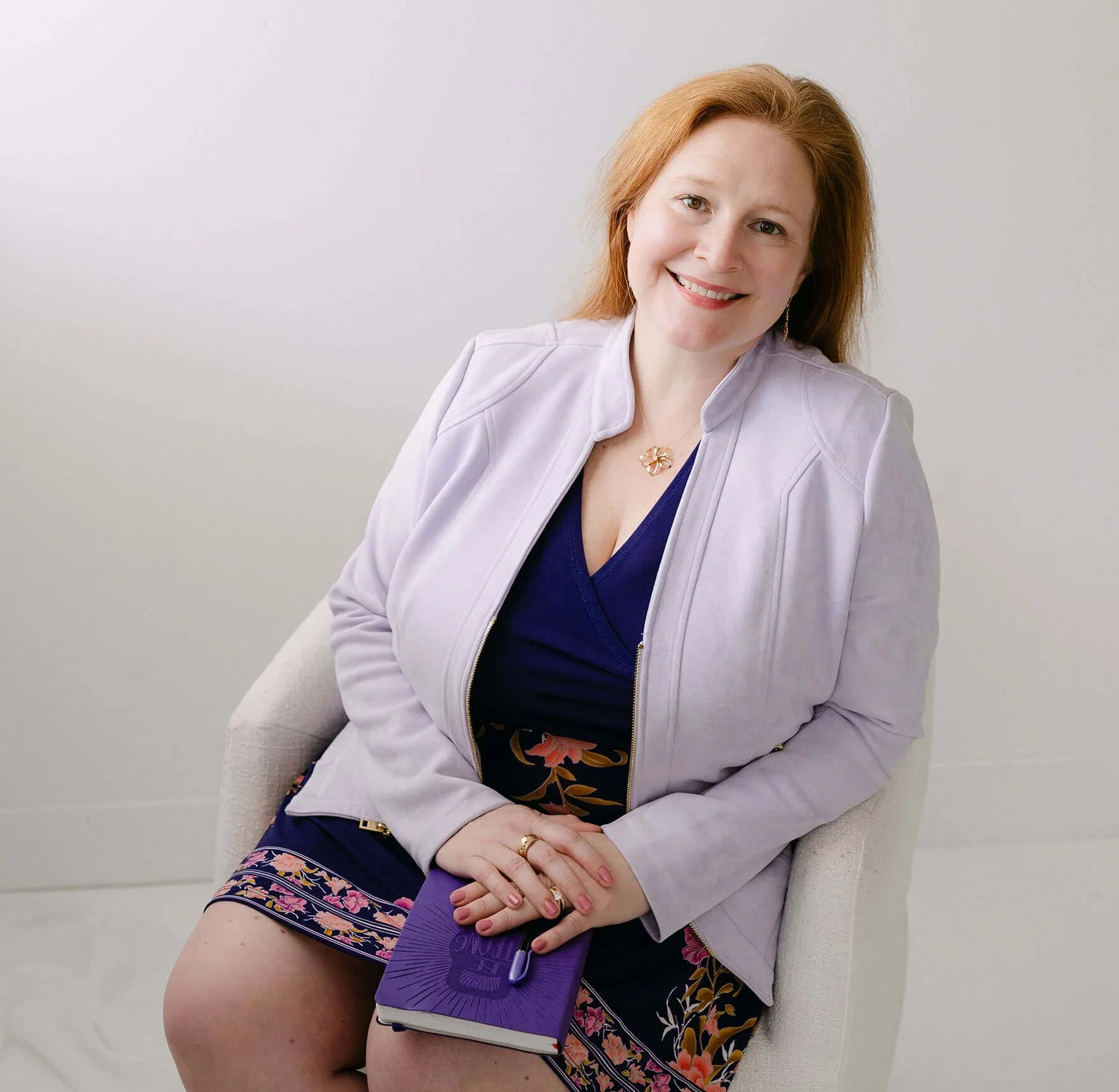 A woman with red hair, wearing a white jacket, dark blue top, and floral skirt, is sitting on a light-colored chair against a plain white background, smiling and holding a purple fan.