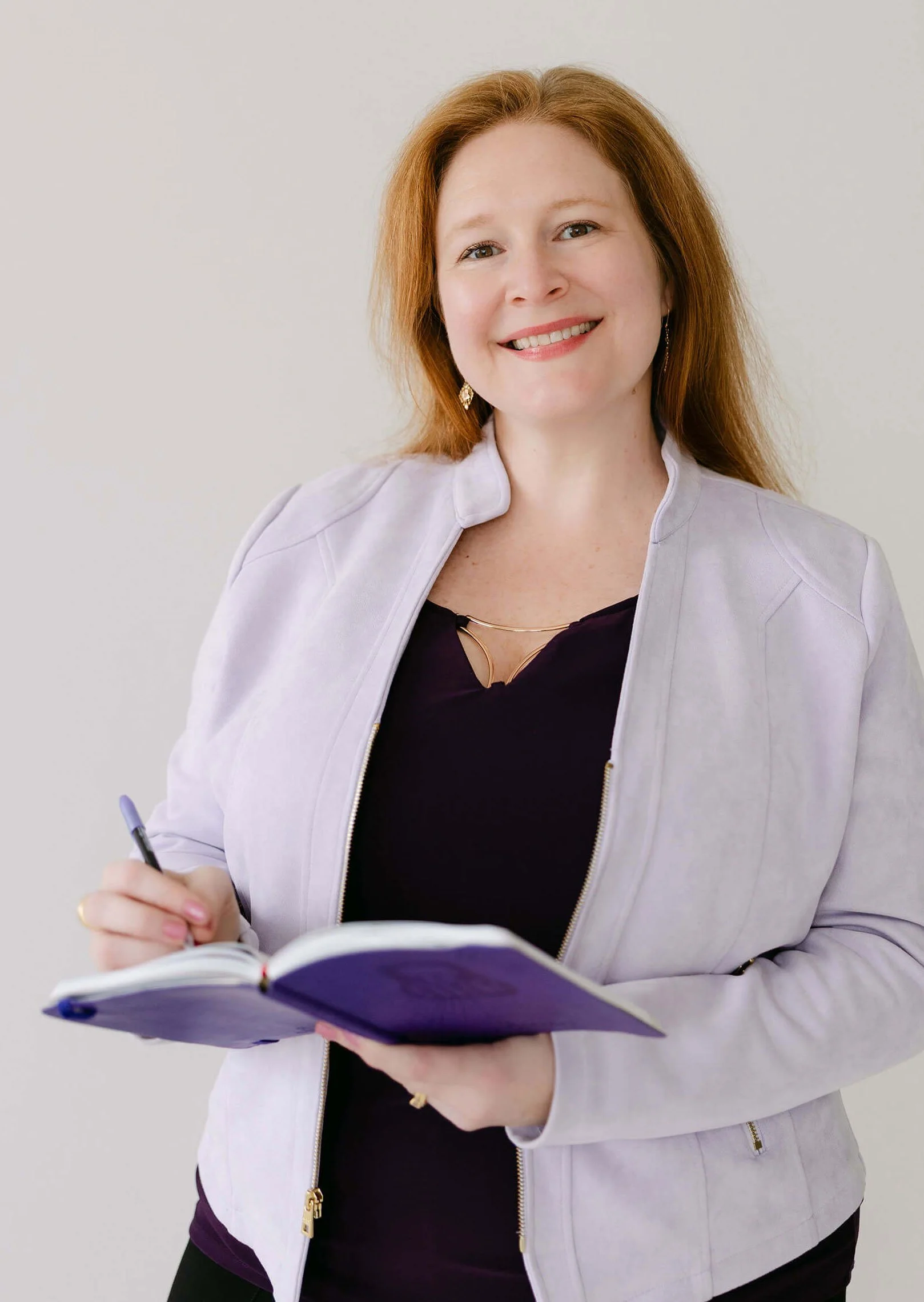 A woman with red hair smiling, holding a pen, and writing in a notebook. She is dressed in a black top and a light-colored jacket, and is standing against a light background.
