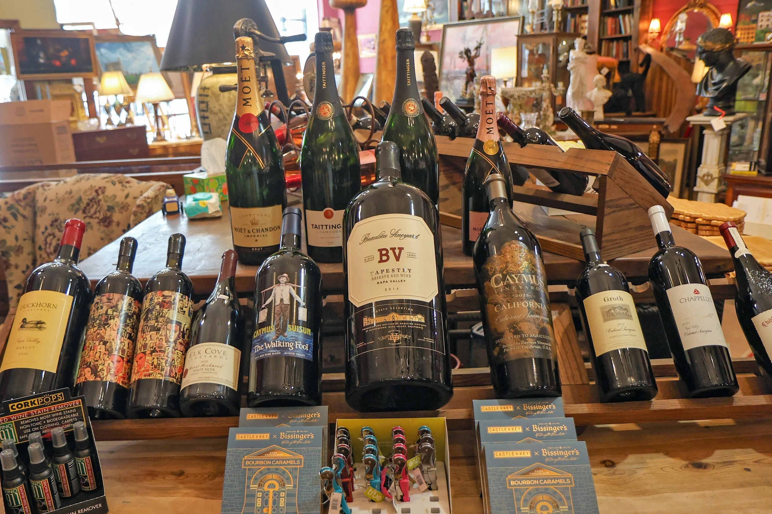 A display of several bottles of wine on a wooden shelf in an antique or thrift store, with various other items like paintings, busts, and lamps in the background.