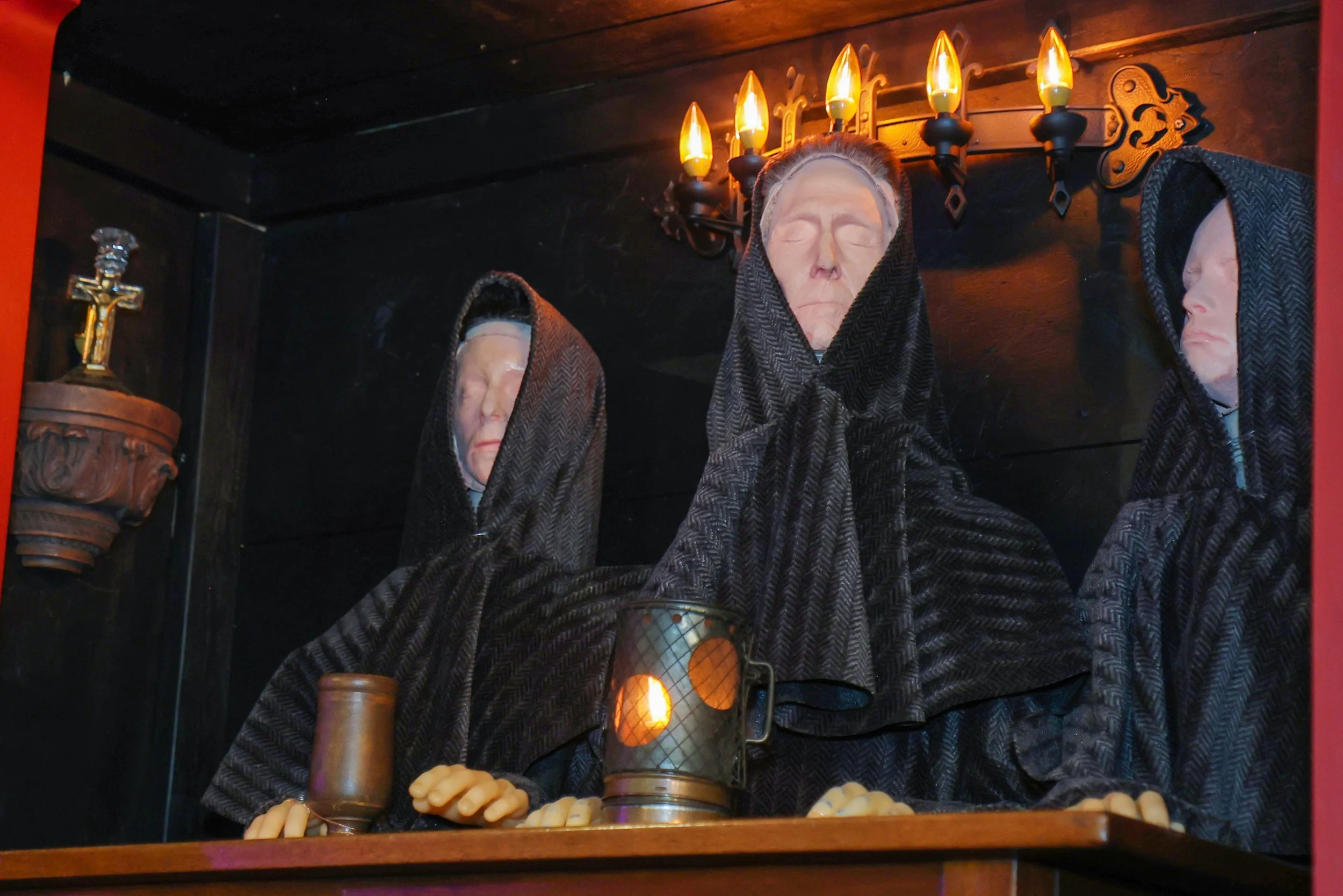 A display of wax figures of elderly people with closed eyes, dressed in dark hoods. The figures are situated under a wall sconce with multiple lit candle-shaped lights. There are additional items including a small crucifix and a candle holder visible on the display.