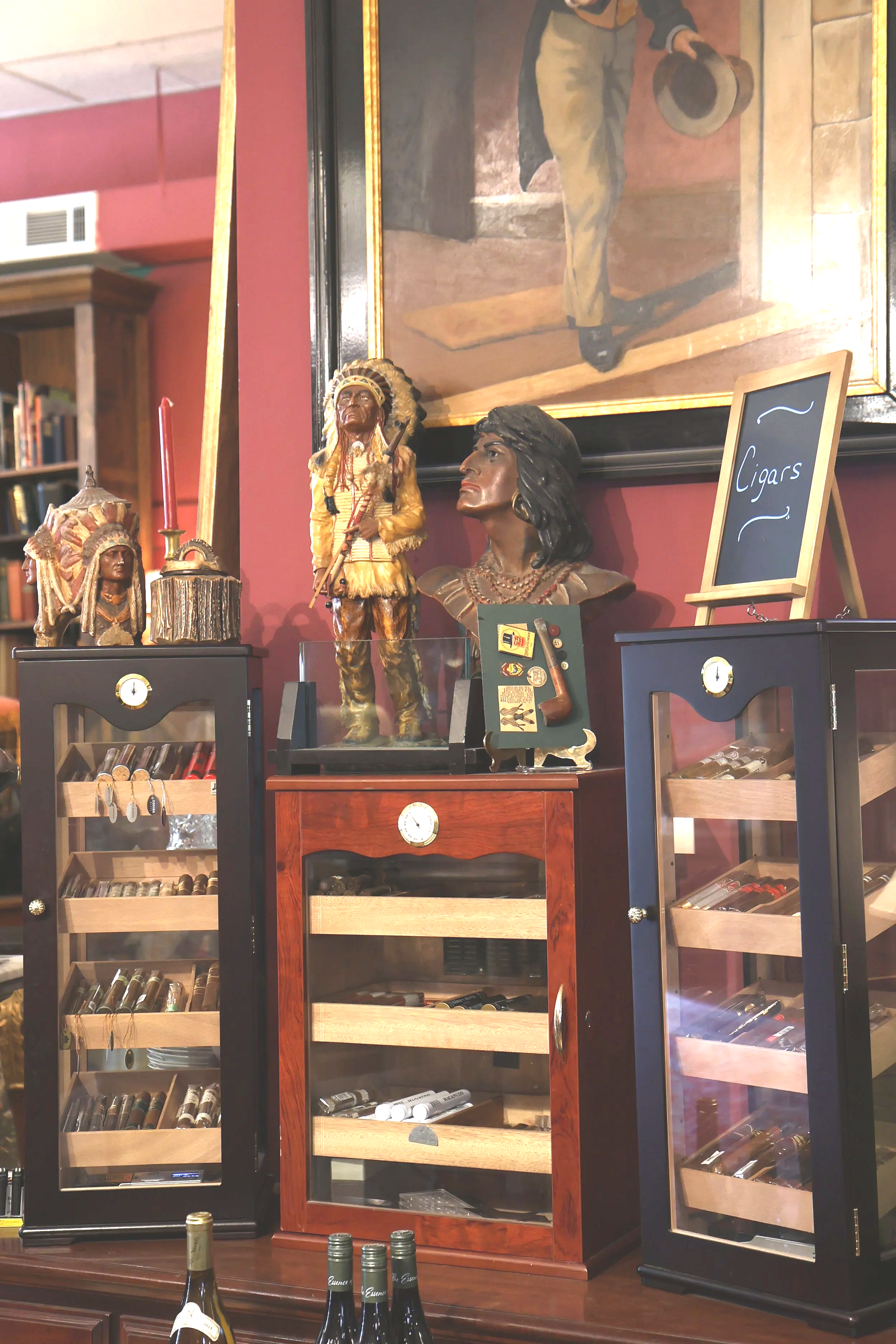Display of cigar humidors with bottles of wine and decorative Native American busts in a shop or bar setting.
