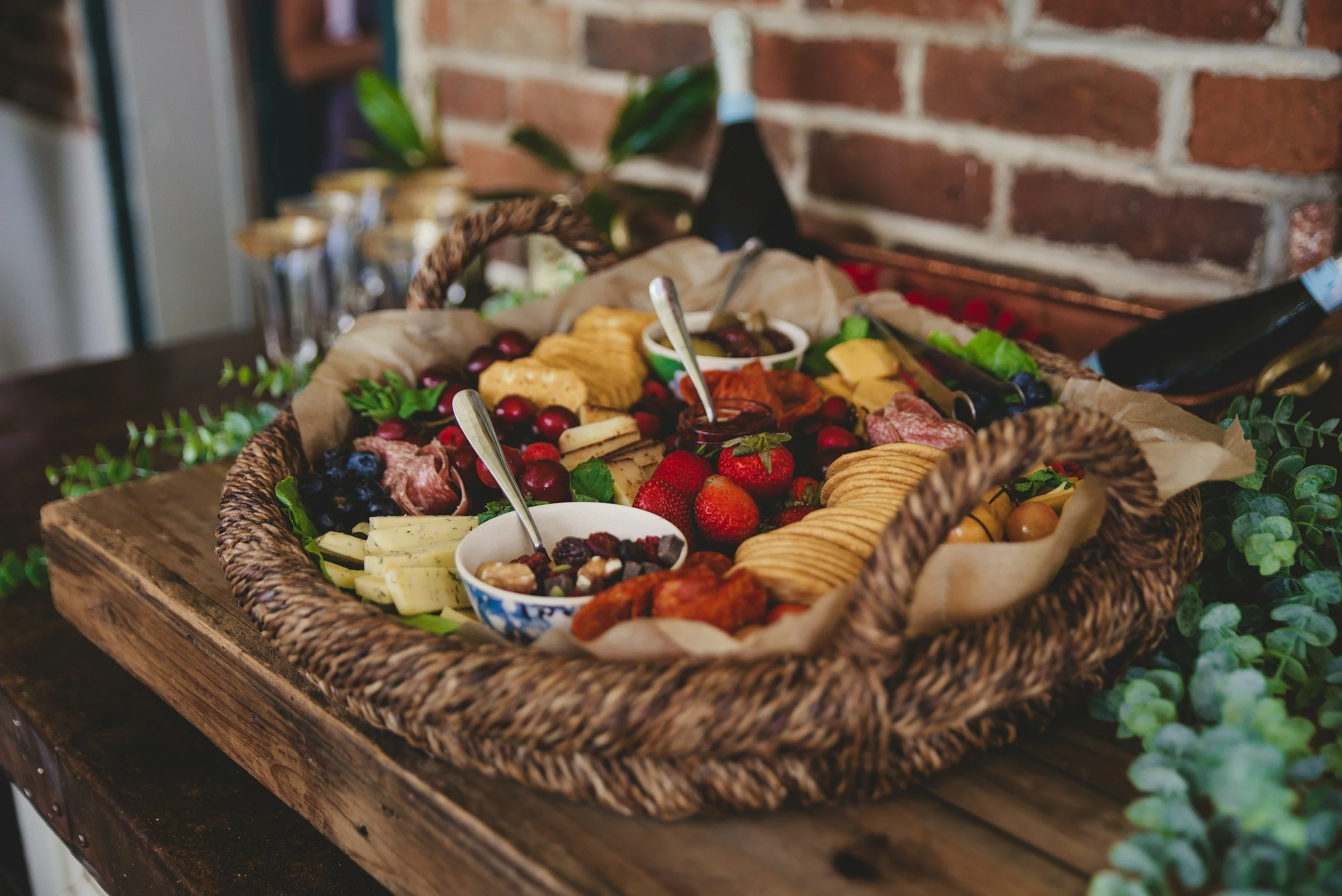 A basket of assorted cheeses, sliced meats, berries, and crackers on a rustic wooden table, with a brick wall in the background.
