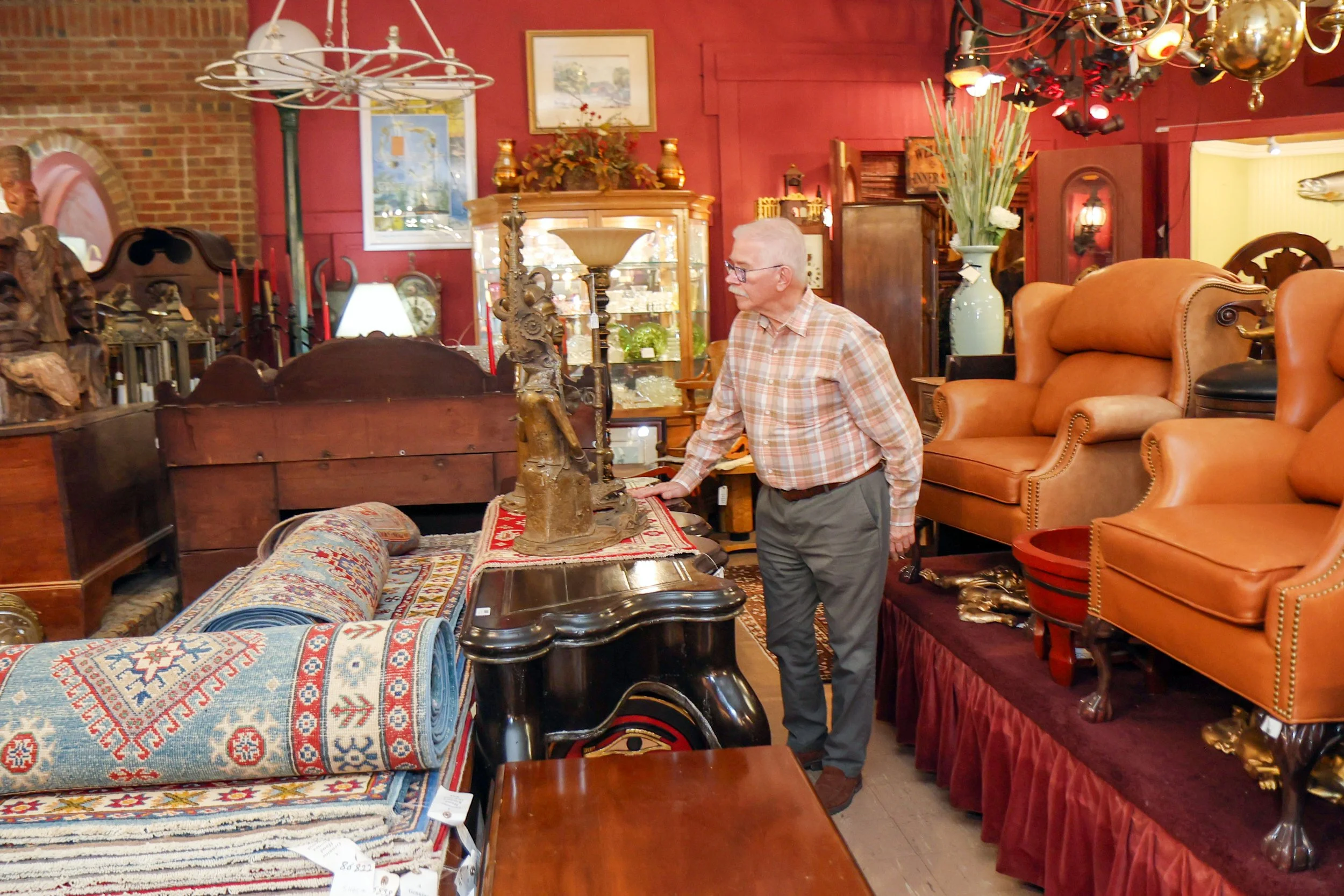 An elderly man with glasses and a mustache is shopping for furniture in a store filled with various sofas, armchairs, and decorative items. The store has red and yellow walls with paintings, vases, and lighting fixtures on display.