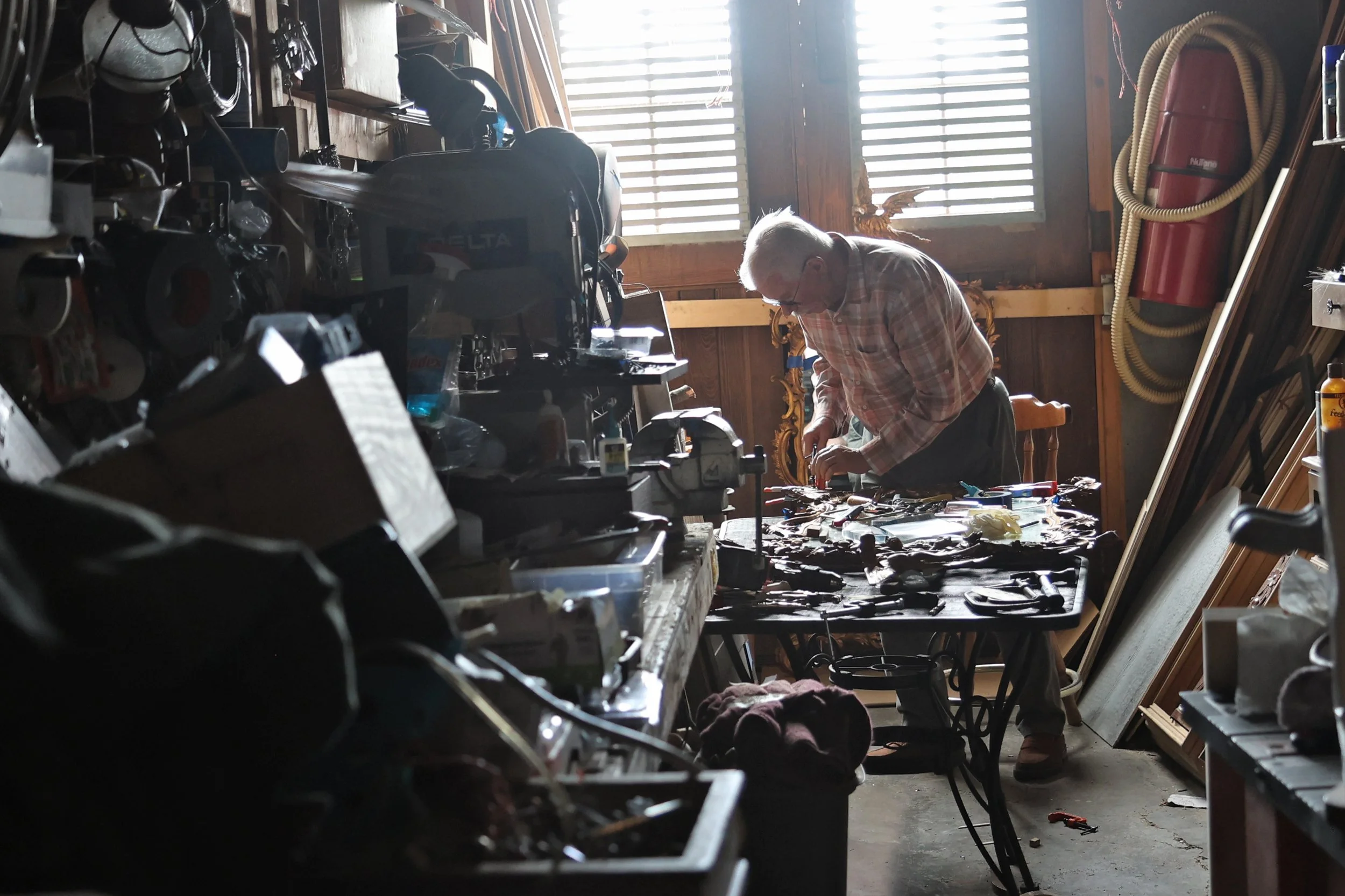 An elderly man with gray hair and glasses working at a cluttered workbench in a workshop, surrounded by tools, equipment, and wooden objects. Light filters through wooden blinds in the background.