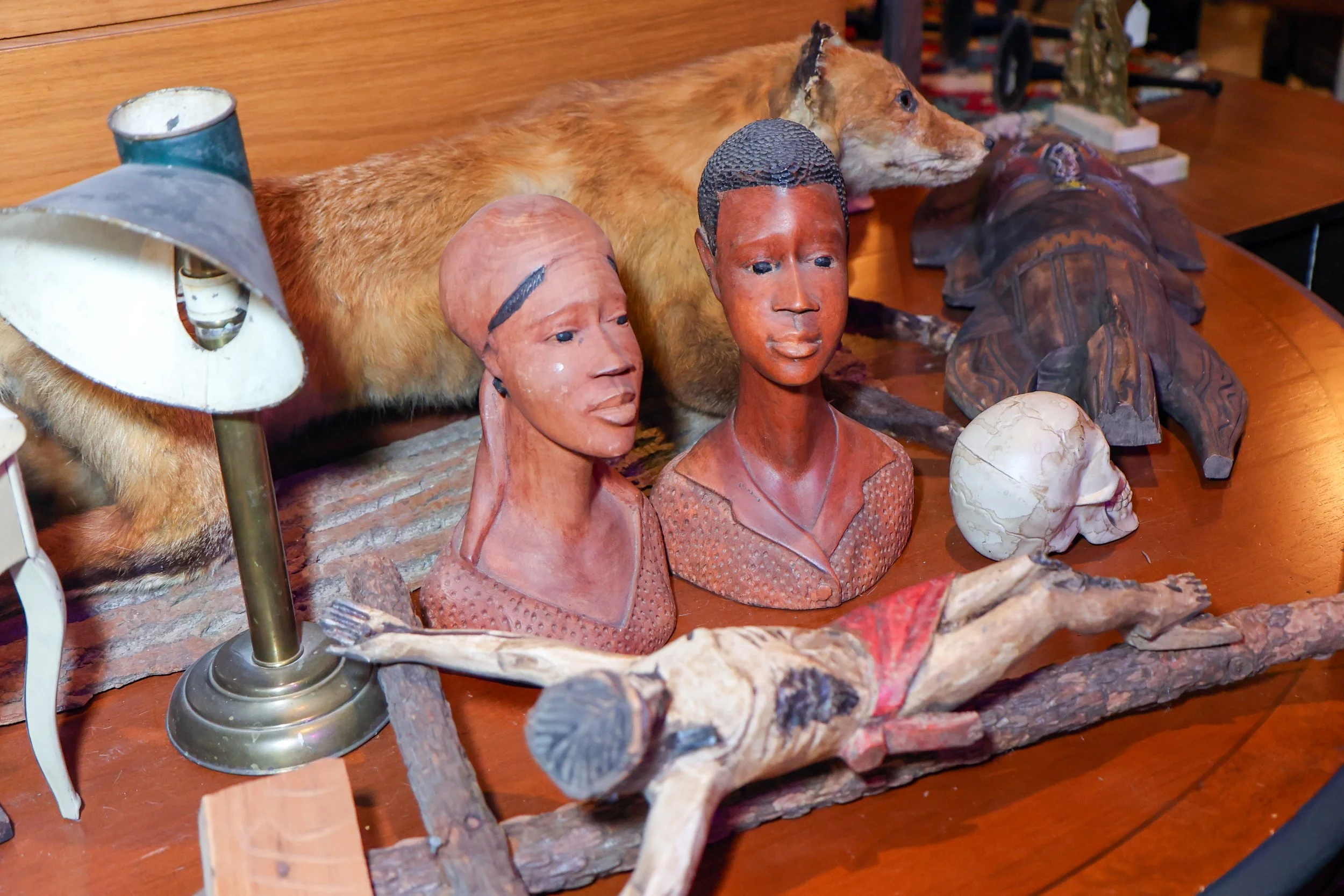 A collection of African tribal art sculptures on a wooden table, including carved busts of men, an animal figure, and masks, with a lamp and other decorative objects in the background.