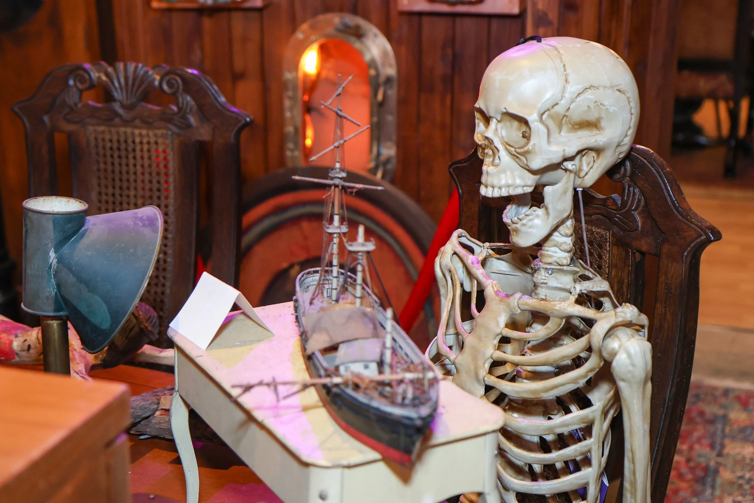A display table with vintage and spooky items, including a human skull on a carved wooden chair, a model boat on the table, a lamp, and various scientific or mechanical objects, in a dimly lit room with wood-paneled walls.