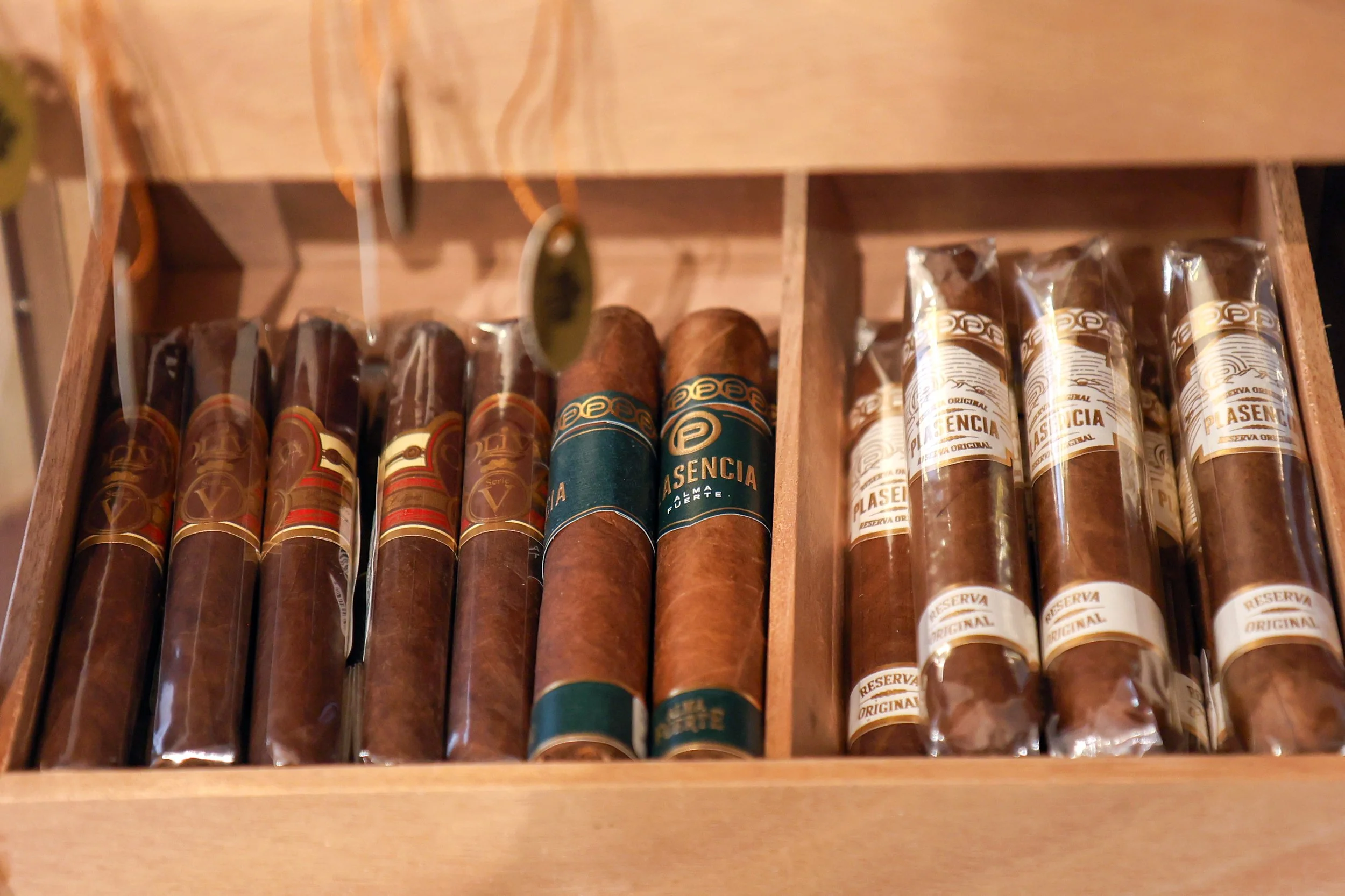 A wooden box containing cigars wrapped in clear plastic, with various labels on each cigar.