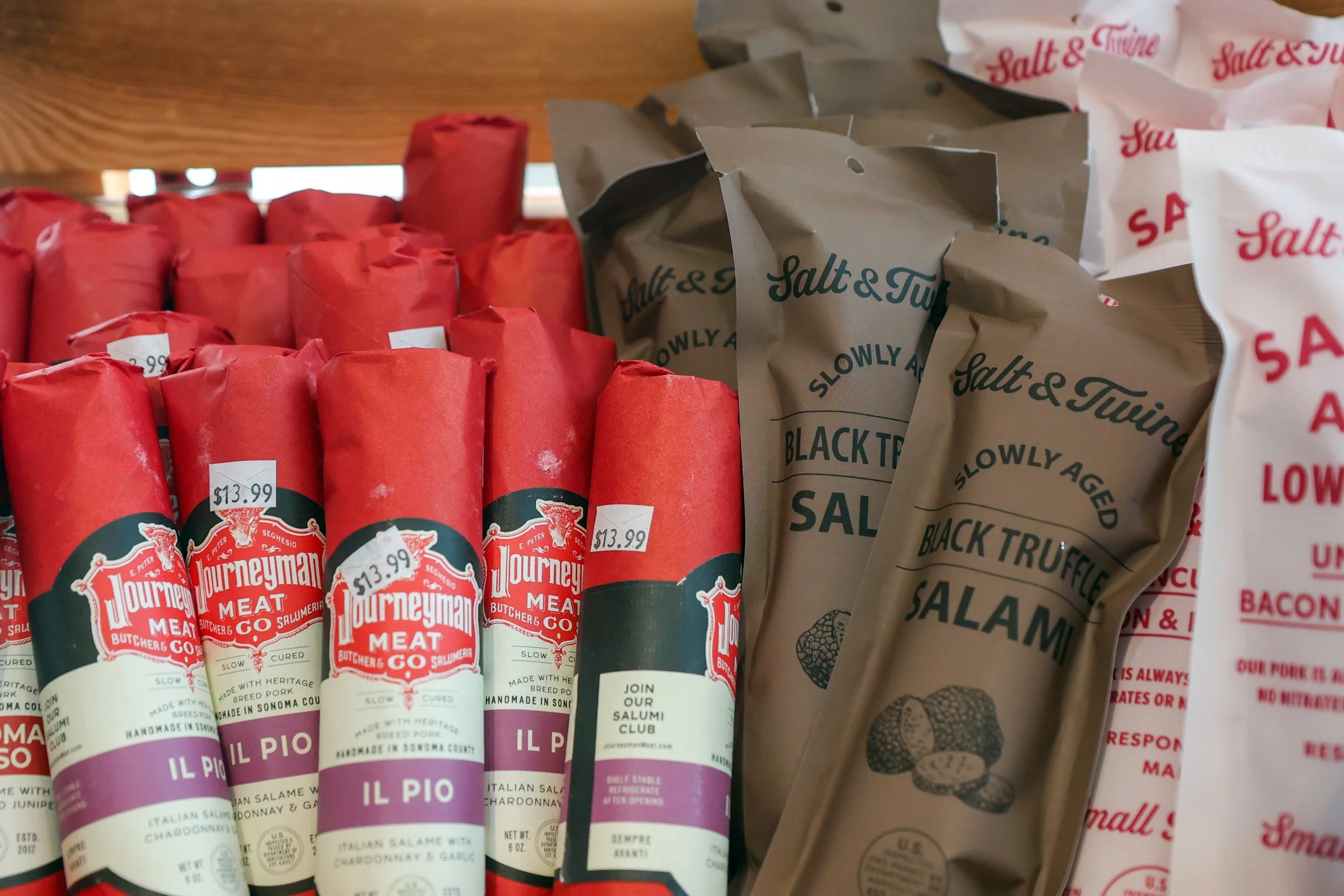 Display of various cured meat sausages and salami packages on a store shelf, with some labeled with prices and brand names.