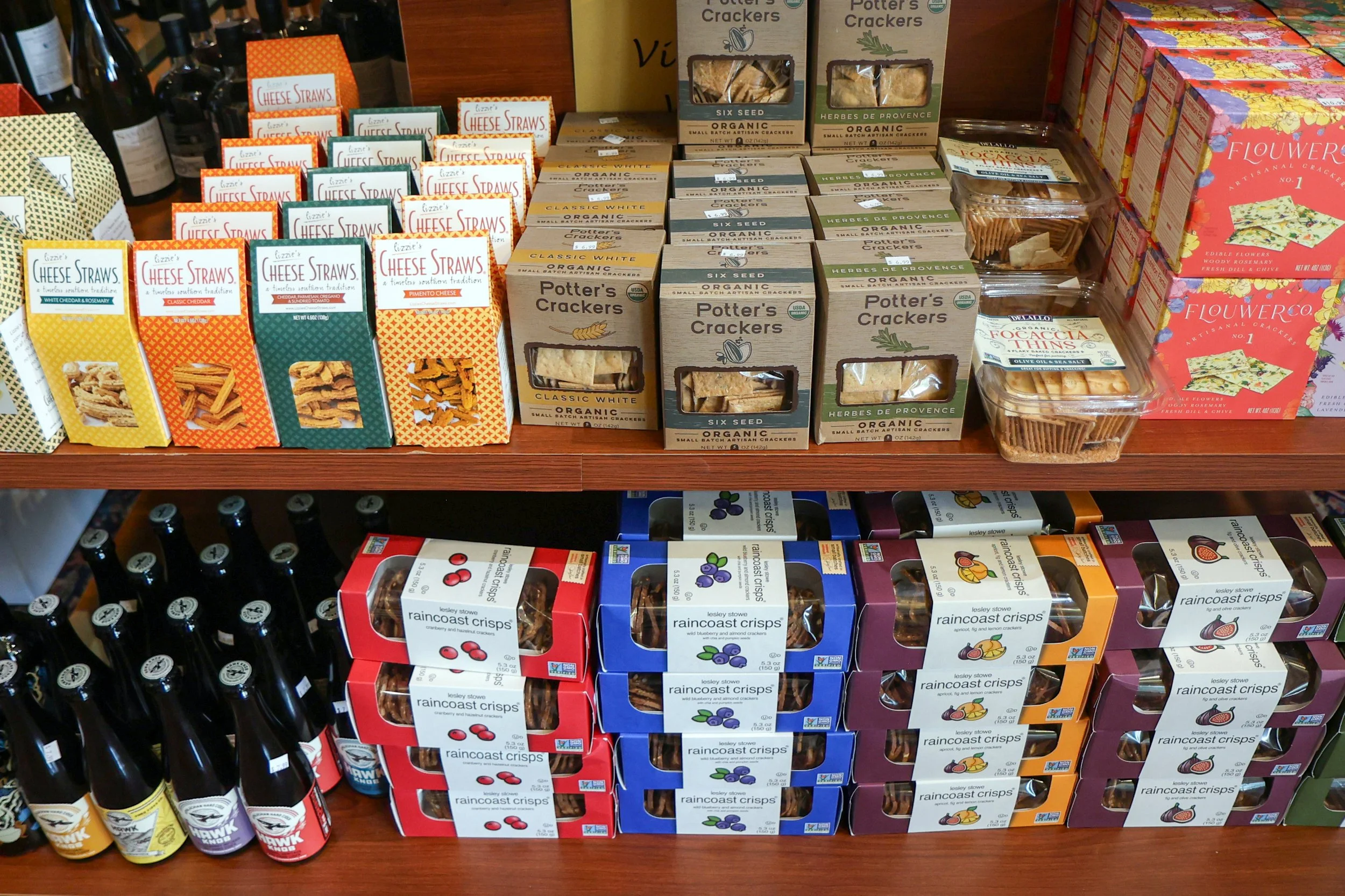 Shelf displaying various snack crackers and crisps including Raincoast Crisps in blueberry, fig and almond flavors, Potters Crackers, and Flower Co. artisan crackers, with wine bottles on the left side.
