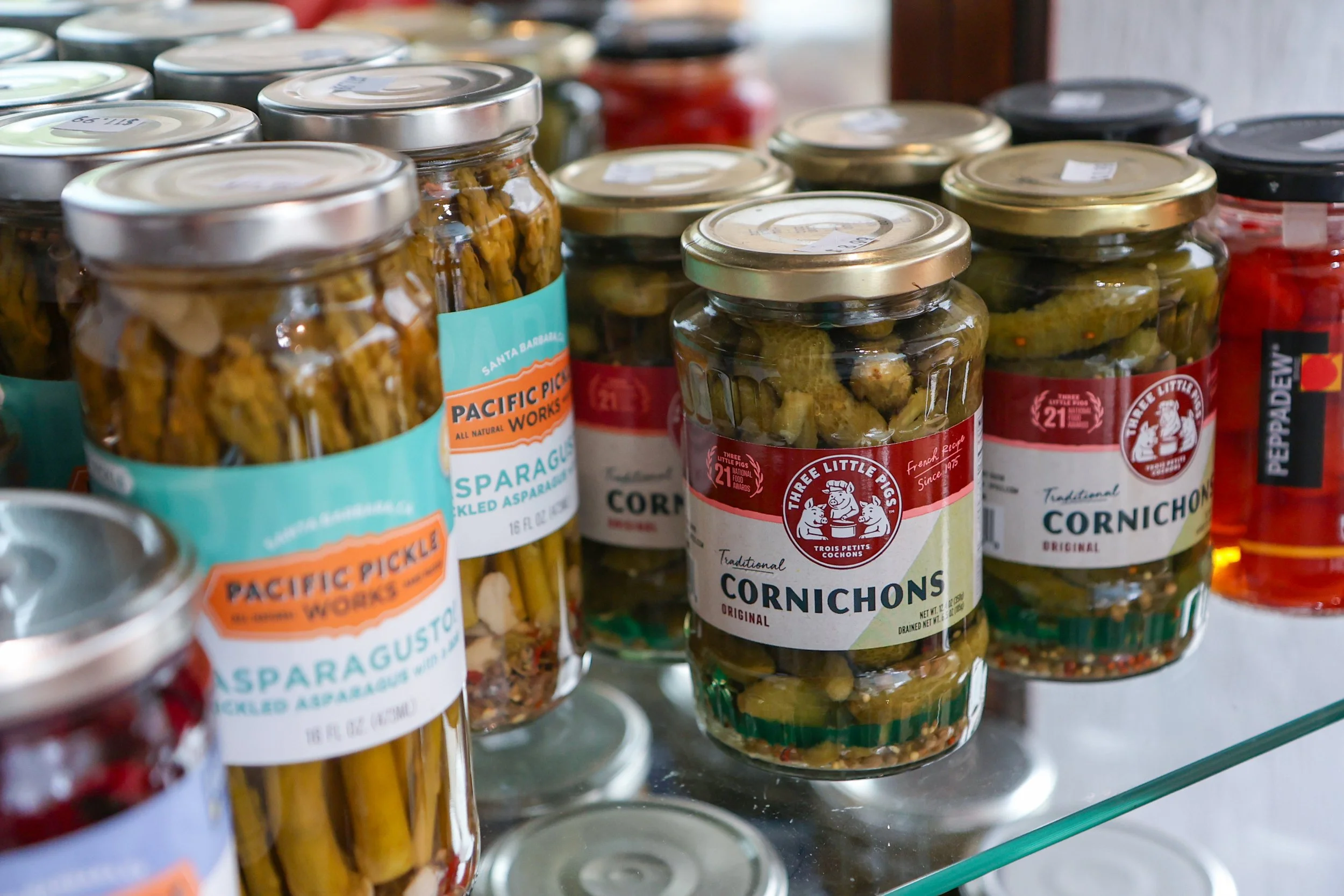 Glass jars filled with pickled vegetables, including asparagus and cornichons, on a store shelf.