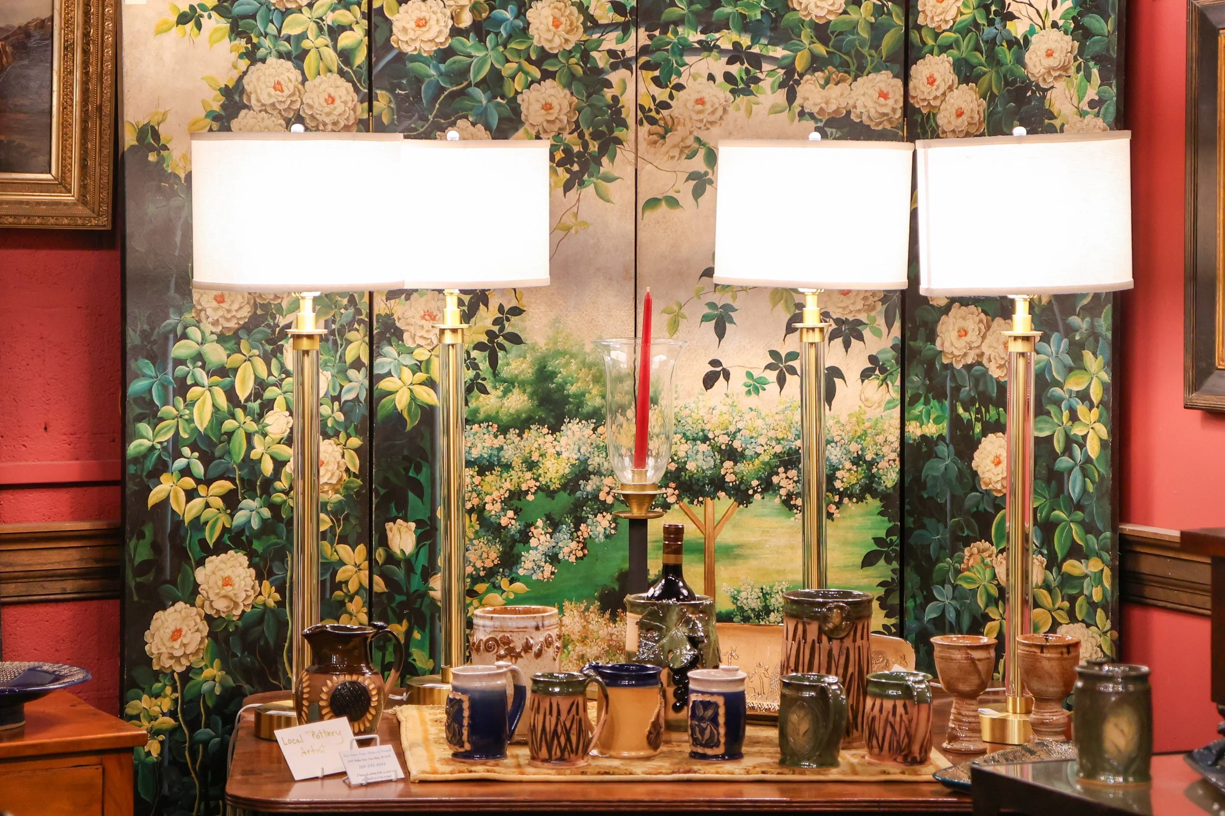 Display of various pottery items on a table with a floral wallpaper background. The pottery includes vases and bowls with decorative patterns. The scene is lit by three tall white lamps with gold bases.