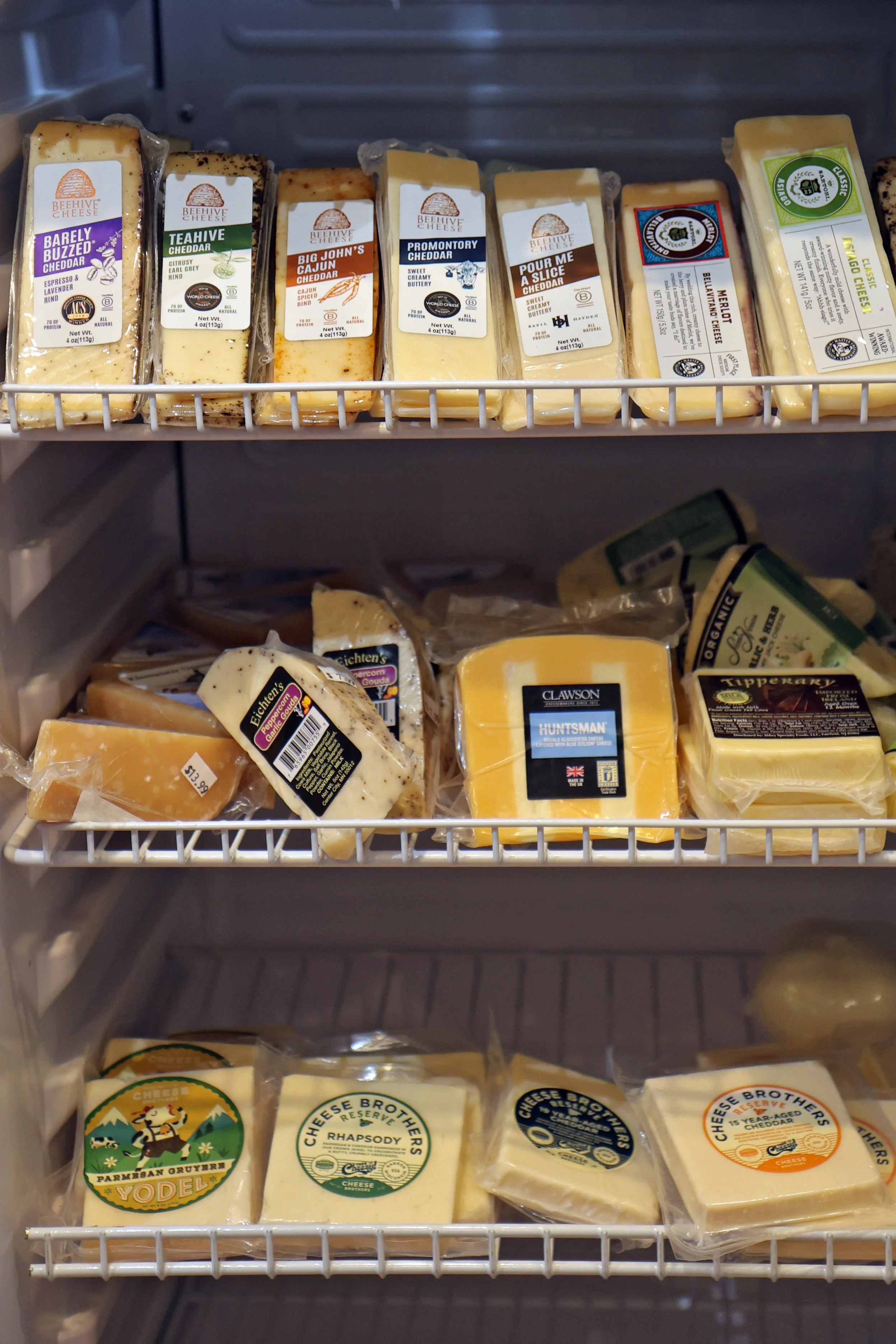 Refrigerator shelf with various blocks of cheese, including cheddar, gouda, and other cheeses from brands like BeeHive, Cheese Brothers, and others.