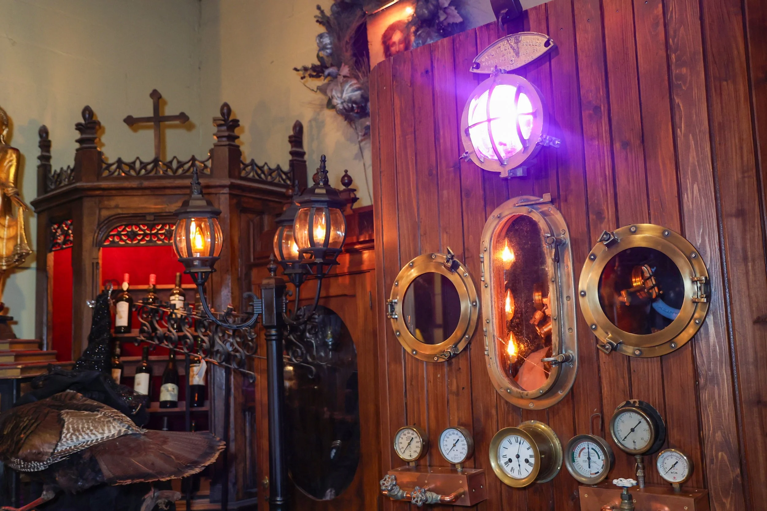 Interior of a room with wooden walls decorated with vintage gauges, lighthouse lamps, and framed religious artwork including a Jesus portrait and a cross. There is a red wine rack with bottles, and some pillows and decorative objects on a leather chair in the foreground.