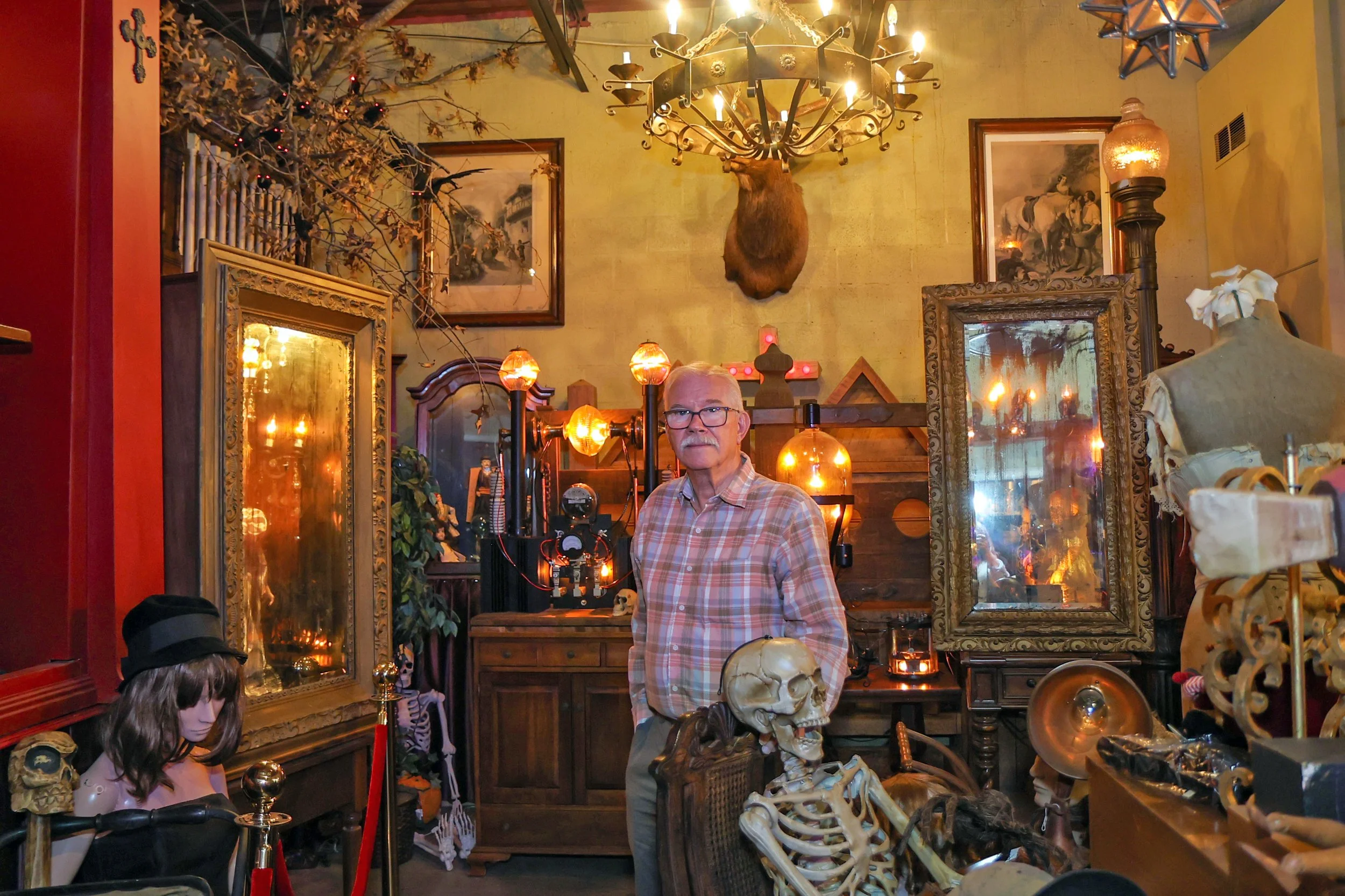 Man with glasses in a decorated room with animal skulls, skeletons, mirrors, and artwork, resembling a vintage or eclectic decor style.