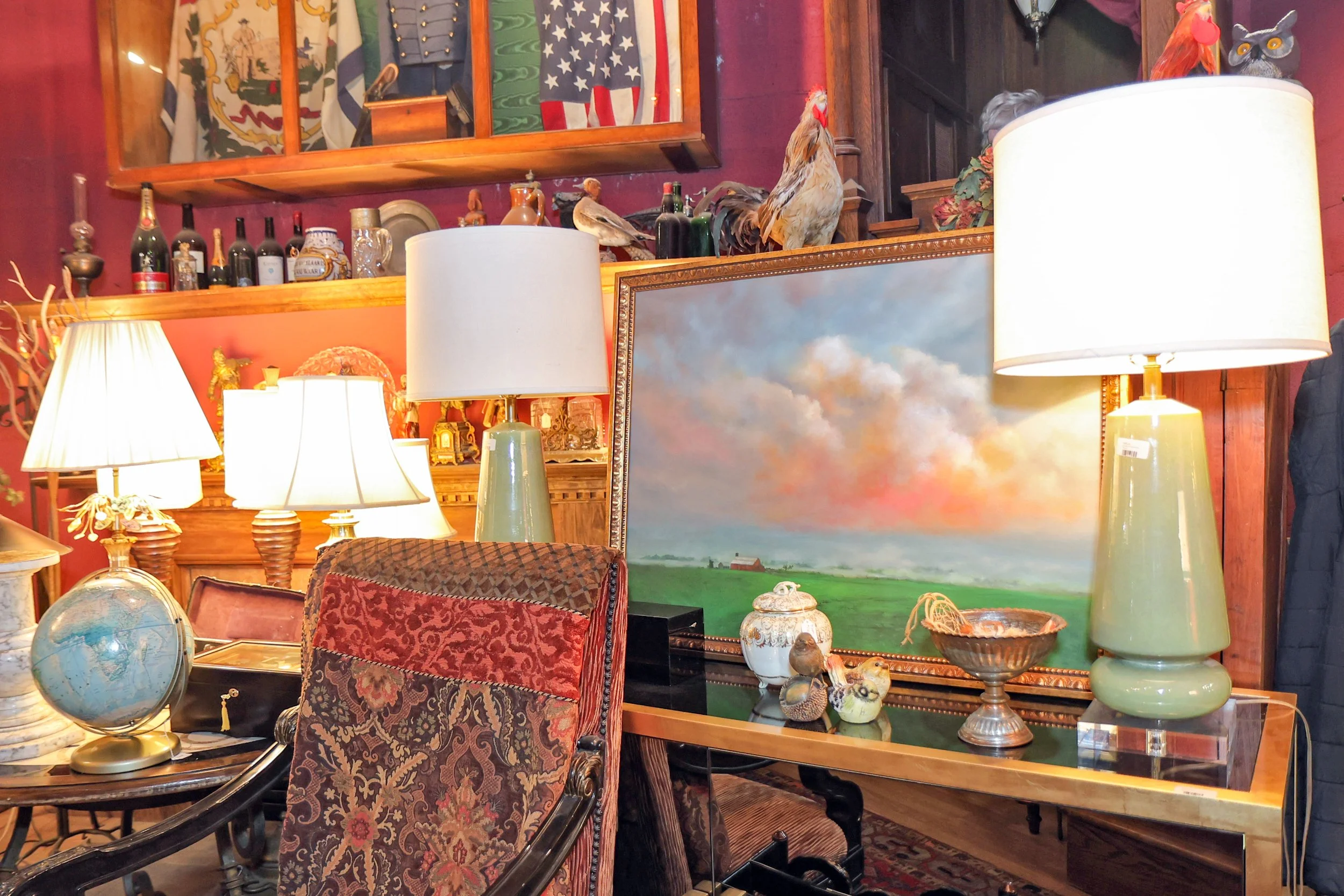 Interior of an antique shop or home decor display featuring lamps, a globe, a painting of a landscape, various decorative objects, and flags on shelves.
