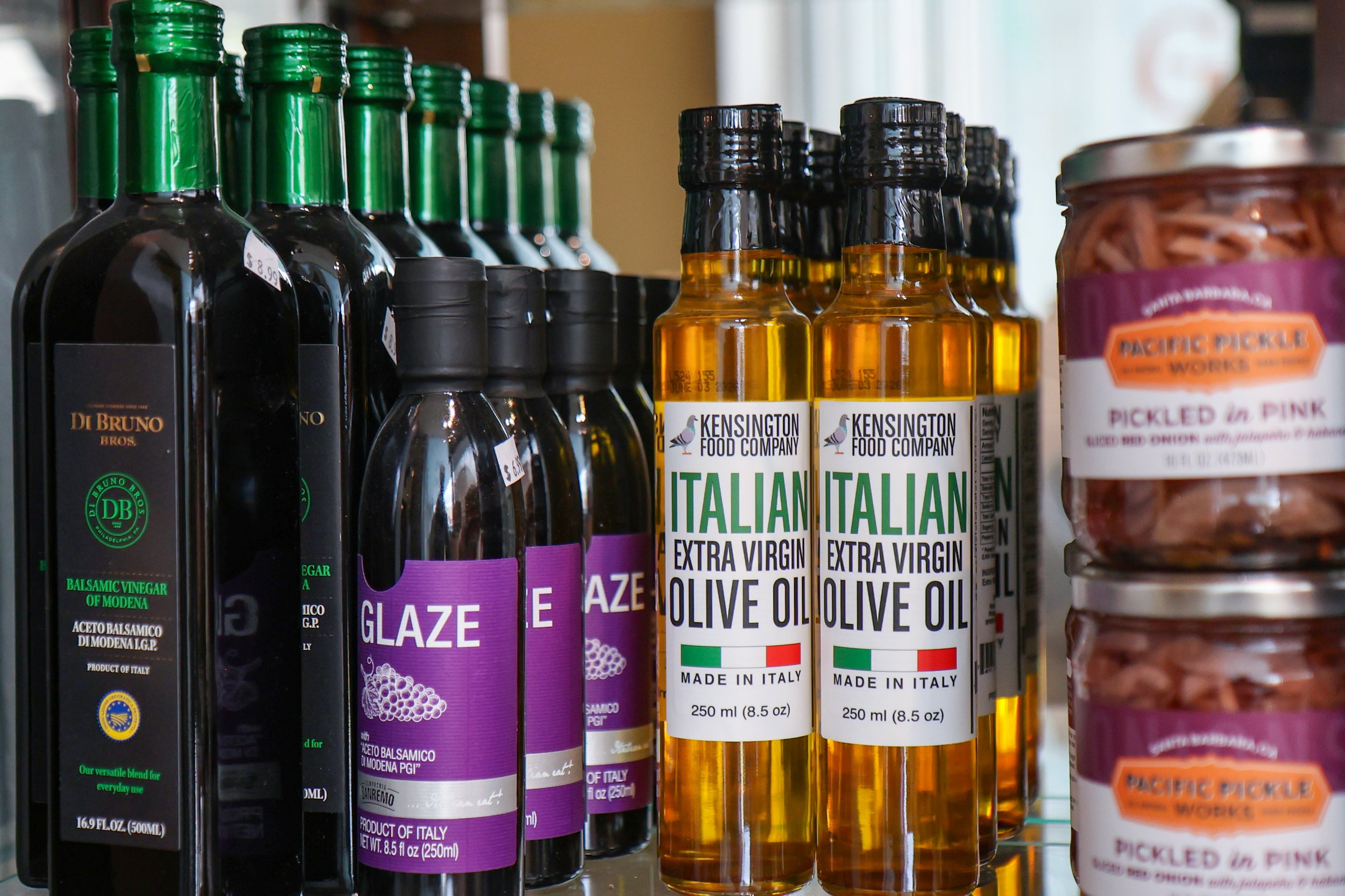 Bottles of balsamic vinegar, olive oil with Italian labels, and a jar of pickled pink ginger on store shelves.