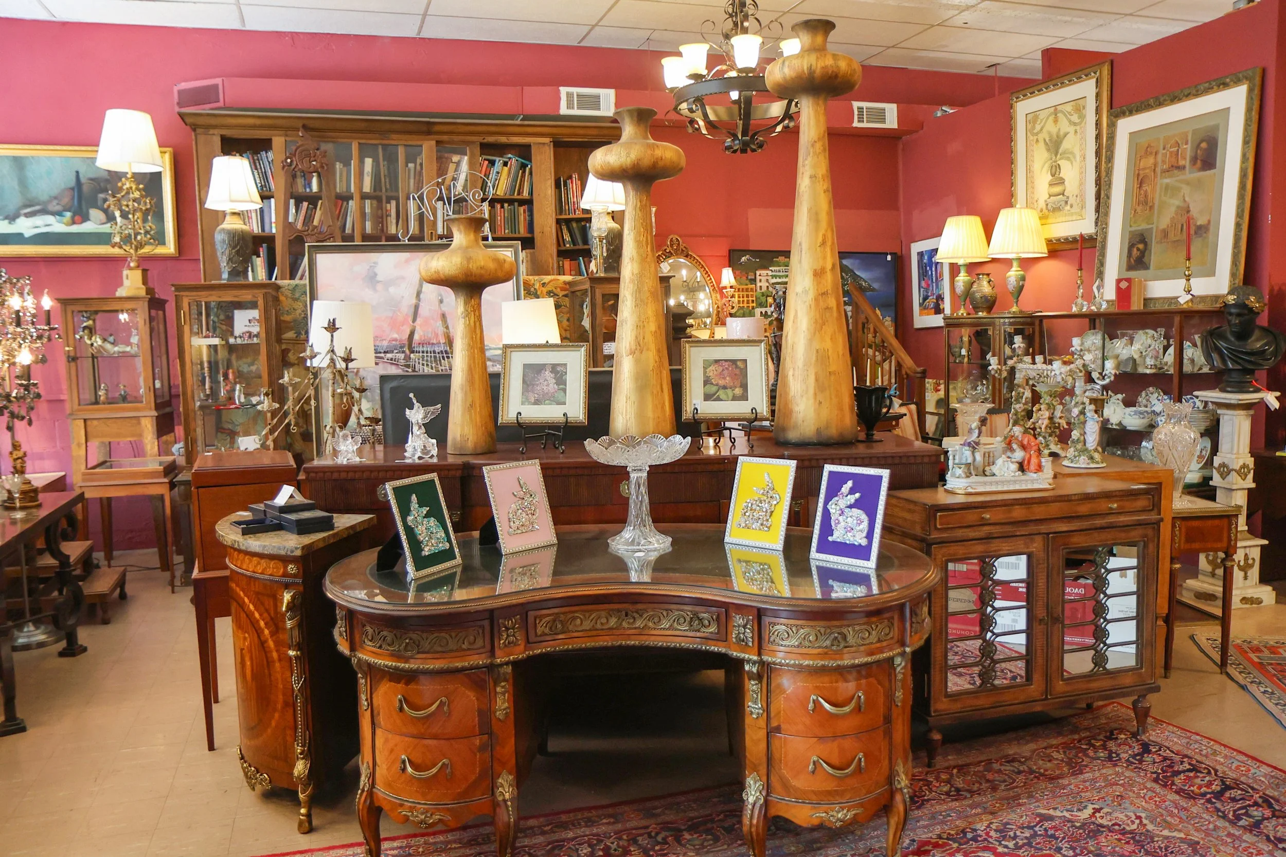 An antique store interior with wooden furniture, lamps, framed artwork, and decorative items, including large wooden vases, framed pictures, and glass figurines, arranged on tables and shelves against red walls.