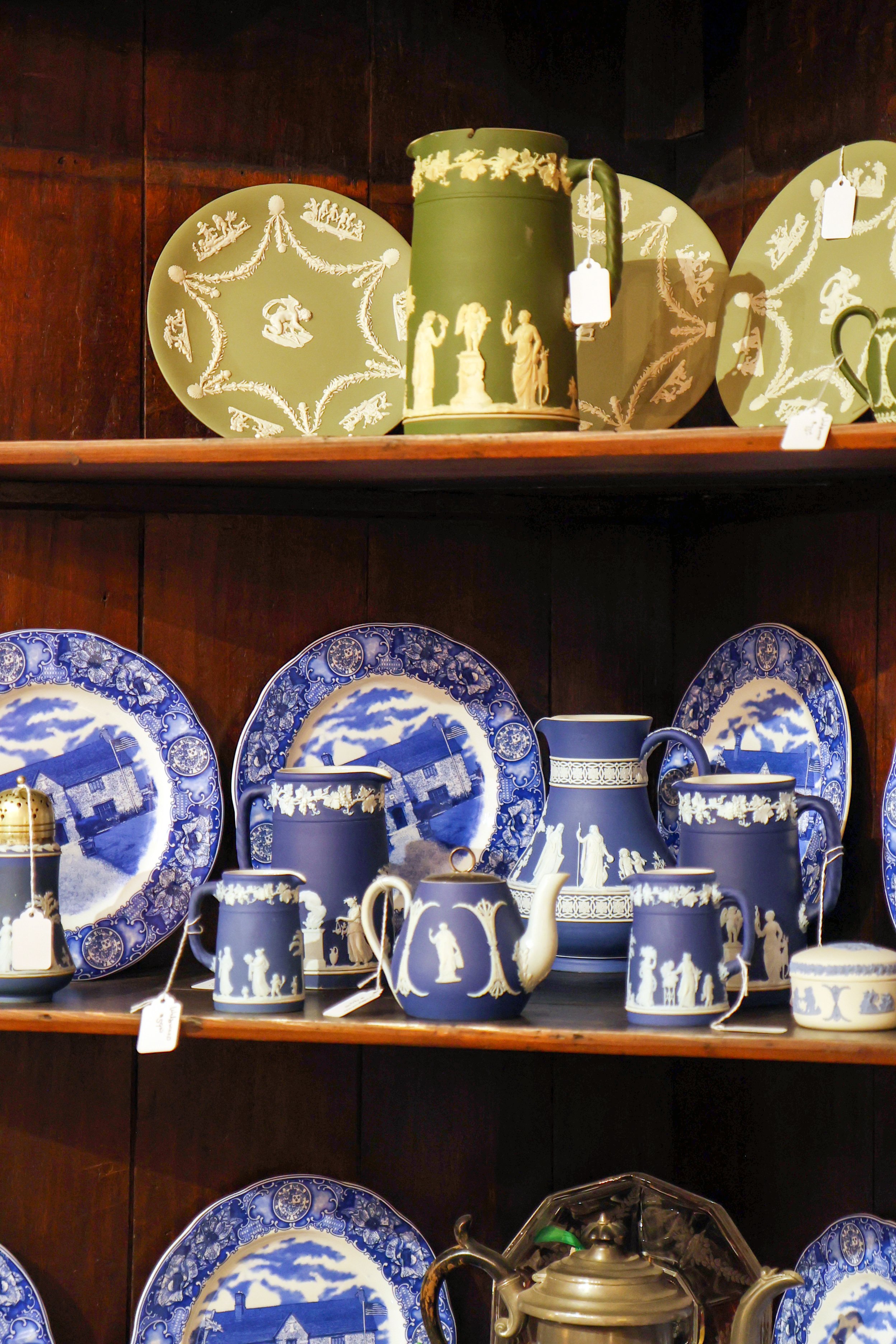 Display of blue and white porcelain plates, cups, and teapots with pastoral and figurative scenes, arranged on shelves.