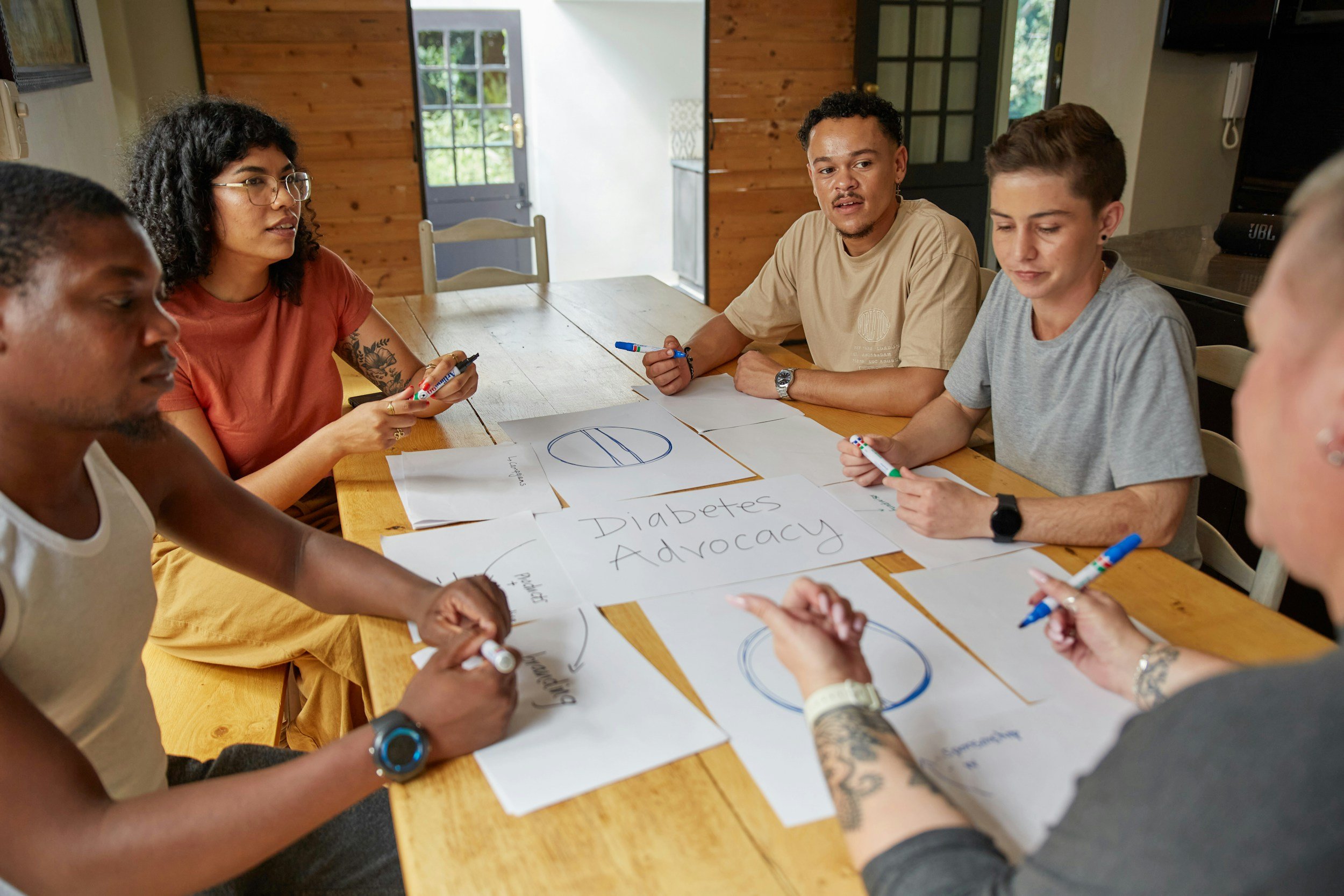 Group of diverse people sitting around a wooden table engaged in a discussion about diabetes advocacy. Papers with diagrams and words are spread across the table.