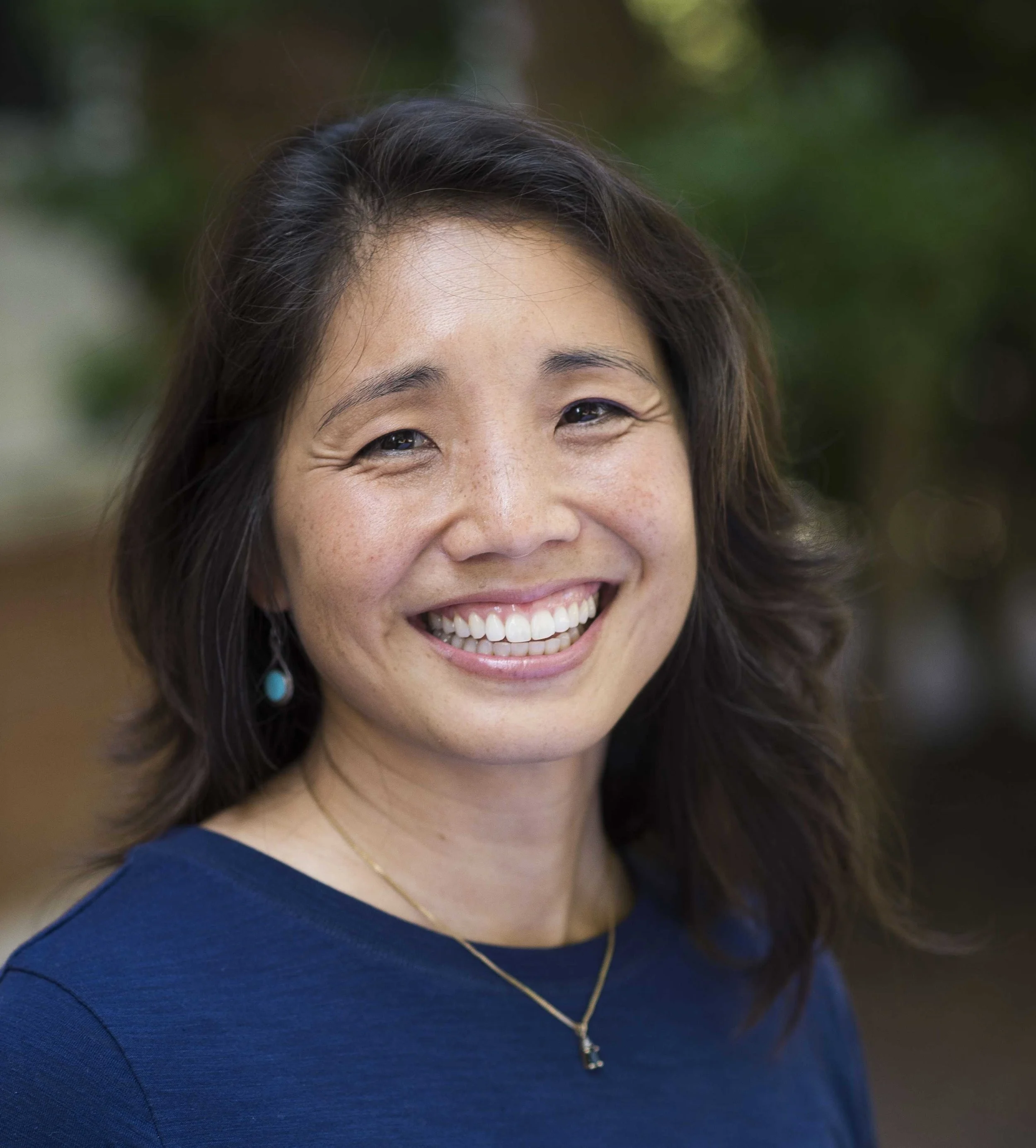 Close-up of a smiling woman with dark brown hair, wearing a dark blue top, gold necklace, and dangling earrings, outdoors with blurred greenery background.