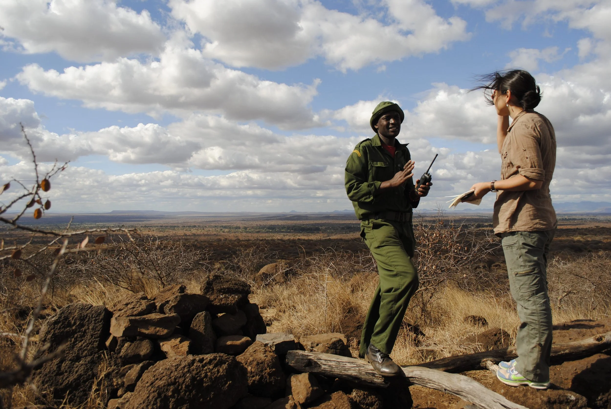 Two women, one in military uniform and the other in casual clothing, are standing outdoors in a landscape with dry grass, rocks, and sparse bushes, under a partly cloudy sky. They appear to be having a conversation, with the woman in uniform holding a walkie-talkie.