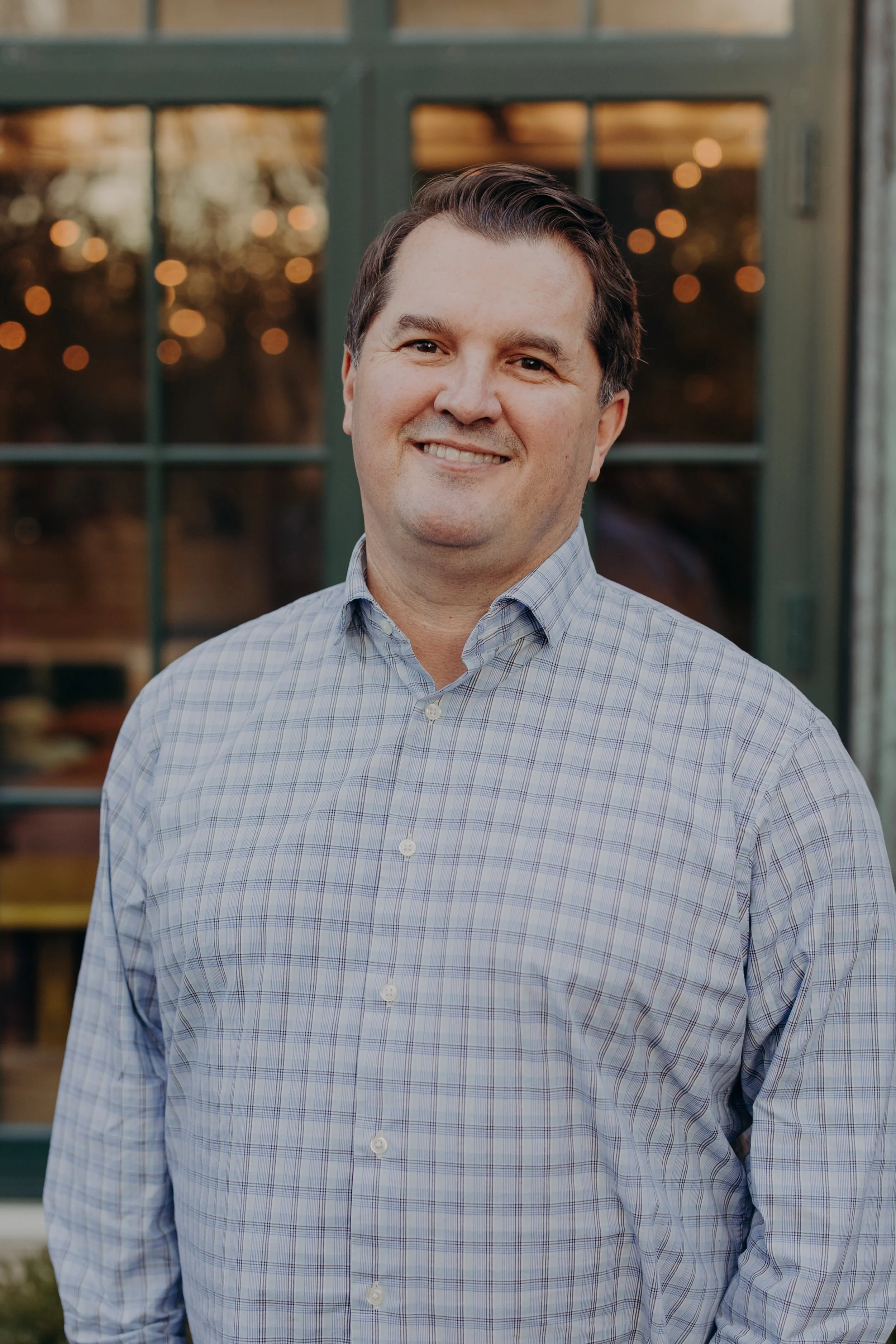 A man in a checked dress shirt standing outside near a window with blurred lights in the background.