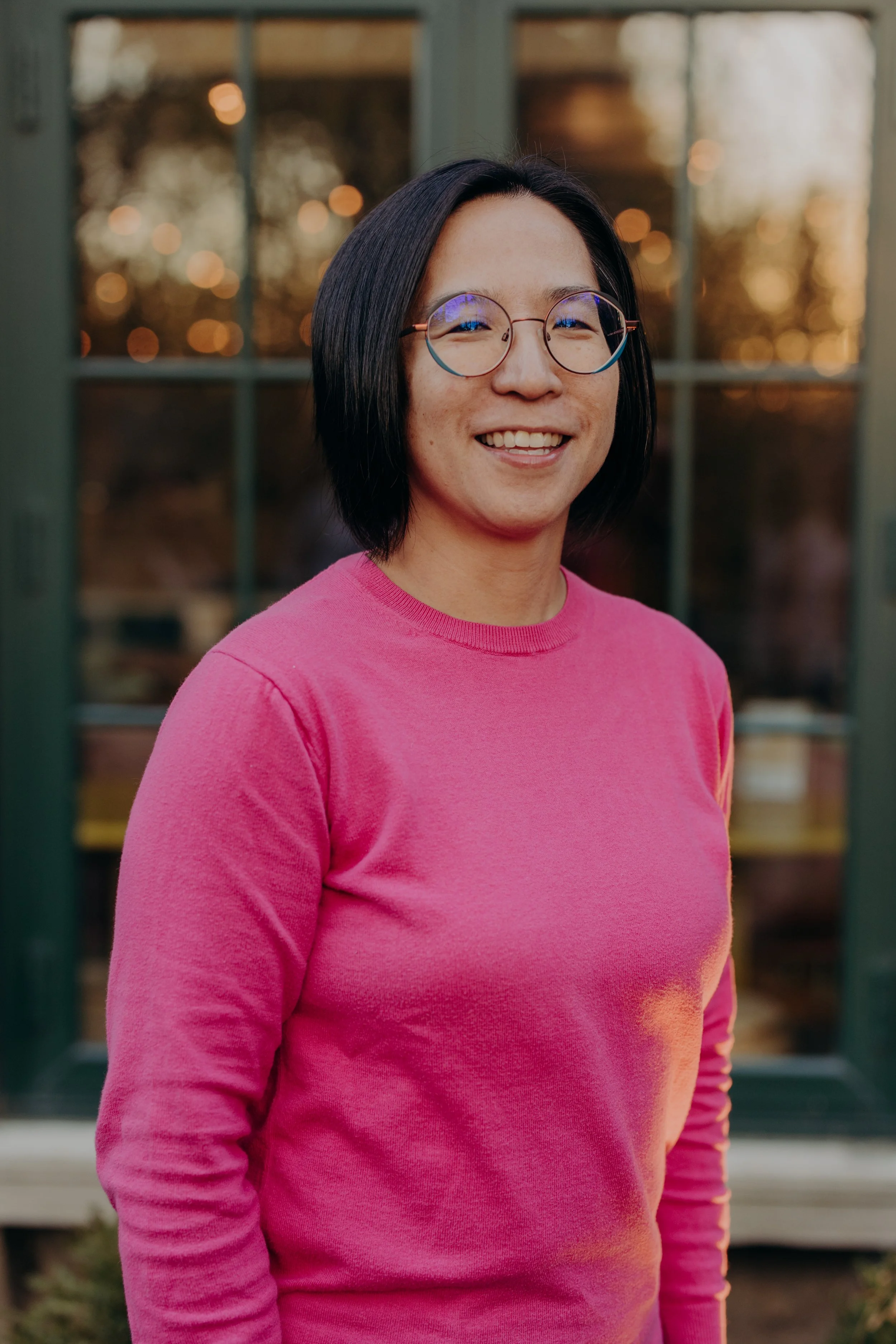 A smiling woman with short black hair wearing glasses and a pink sweater standing outdoors in front of a building with large windows and warm lighting.
