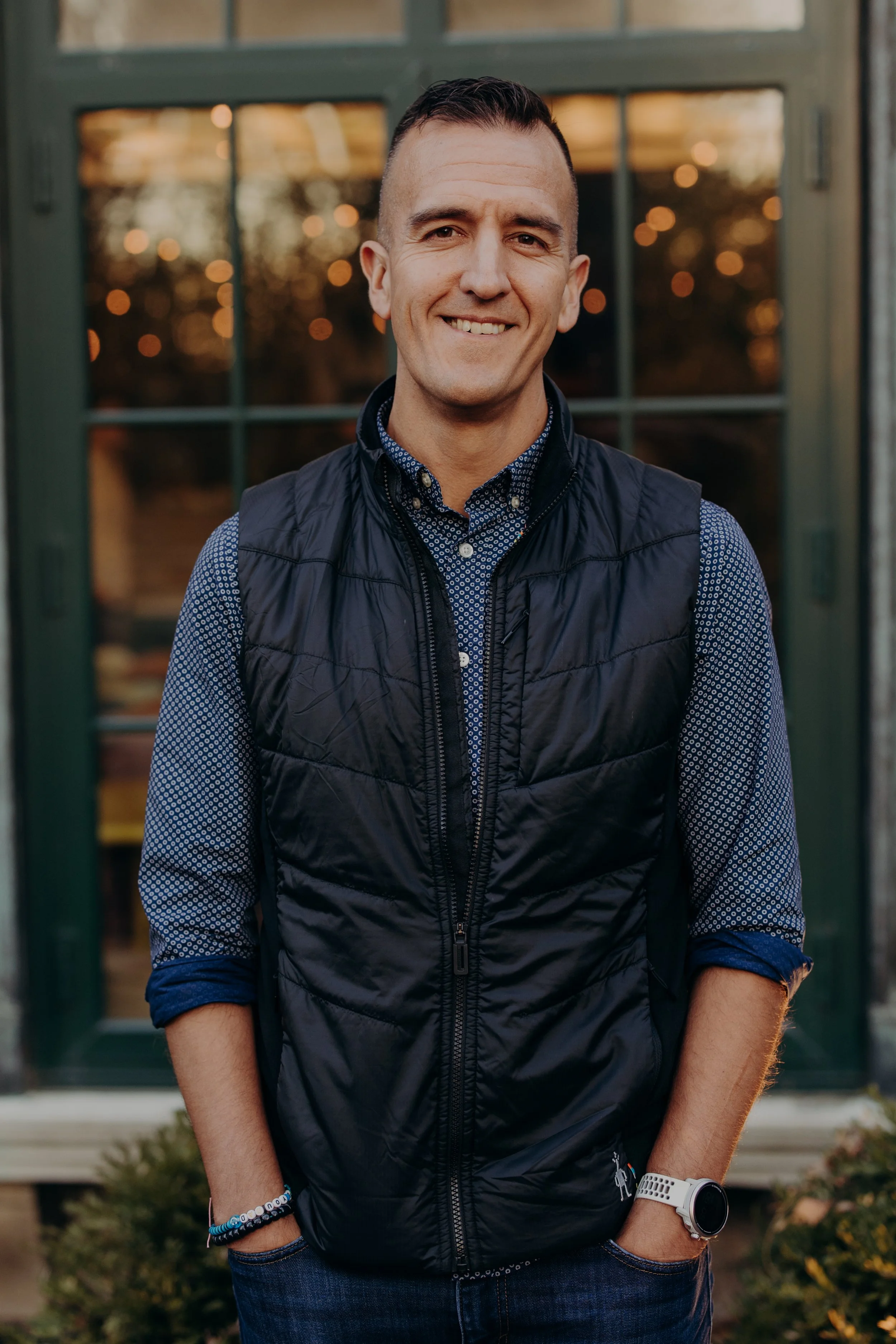 A young man with short dark hair, wearing a blue patterned button-up shirt, a black puffer vest, jeans, and a white sports watch, standing in front of a green-framed window with a warm outdoor setting in the background.