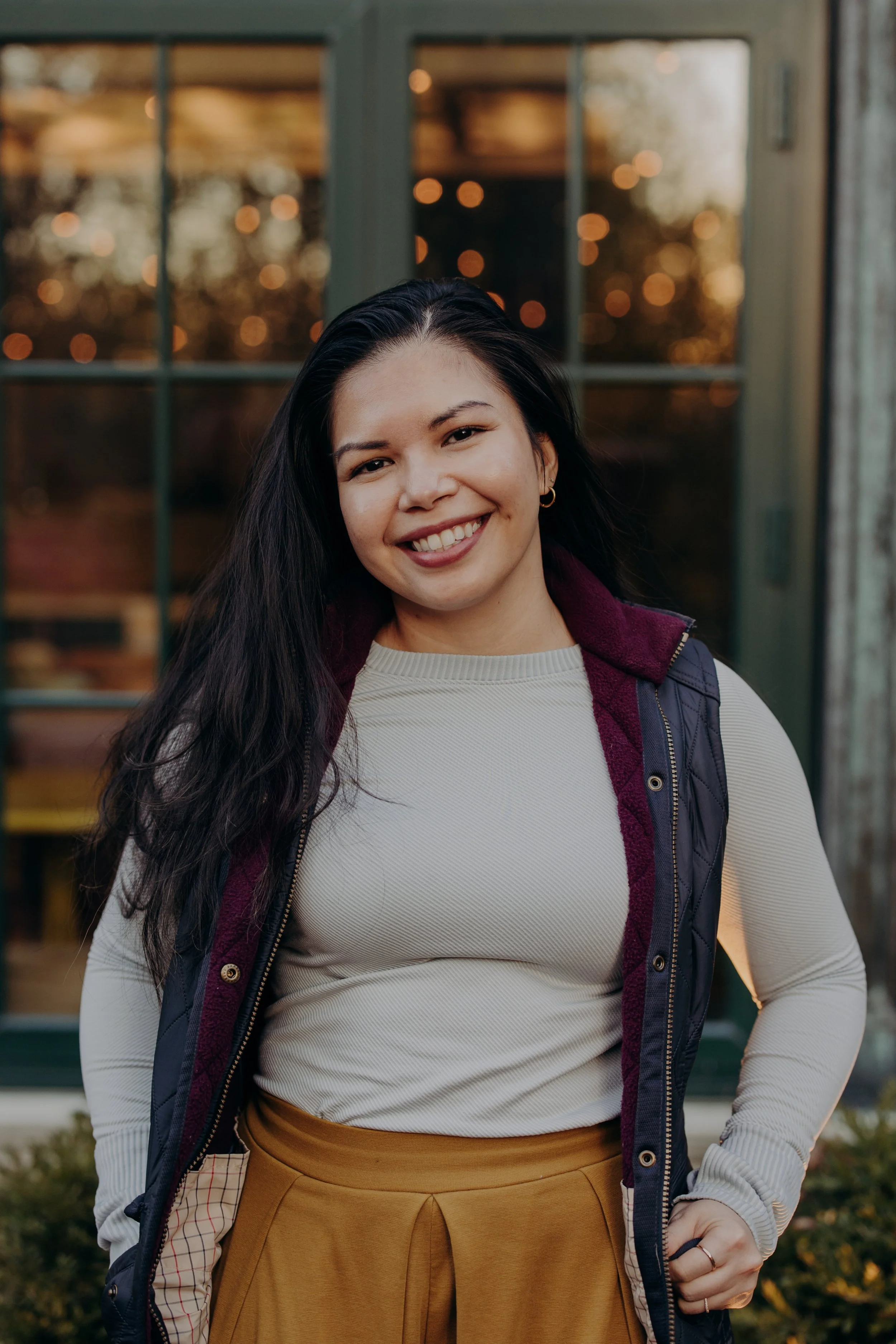 A woman with long dark hair smiling outdoors in front of a window with blurred lights at dusk. She is wearing a light gray long-sleeve shirt, a dark vest with maroon accents, and mustard-colored pants.