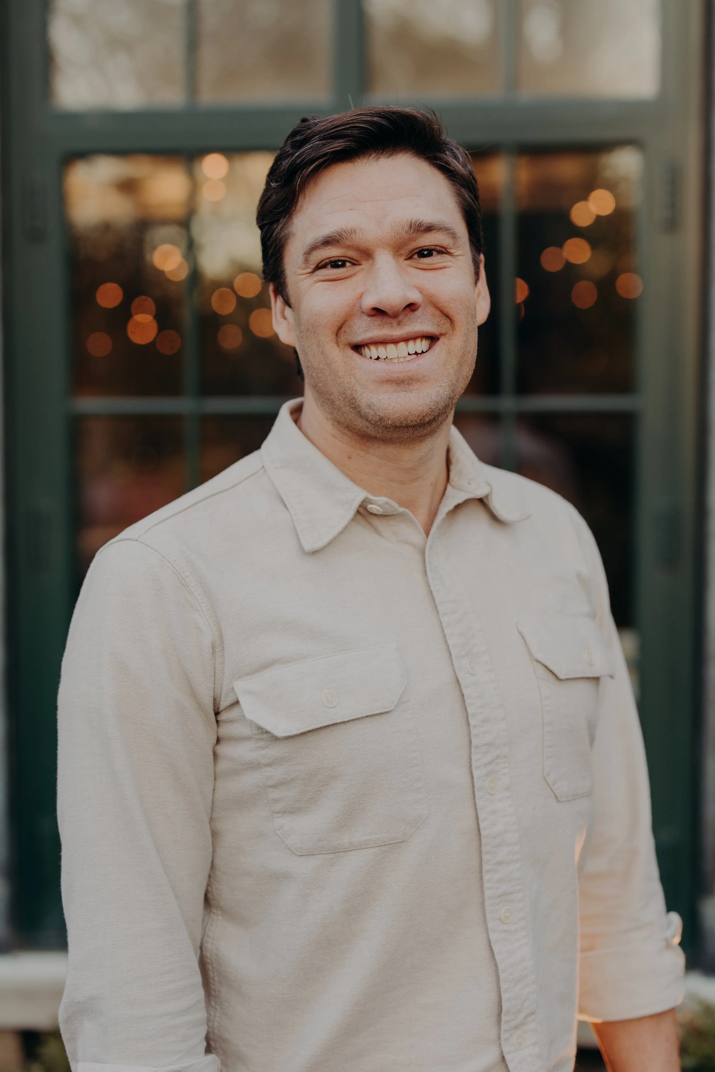 A smiling man with dark hair, wearing a light-colored shirt, standing outside in front of a window with blurry lights behind him.
