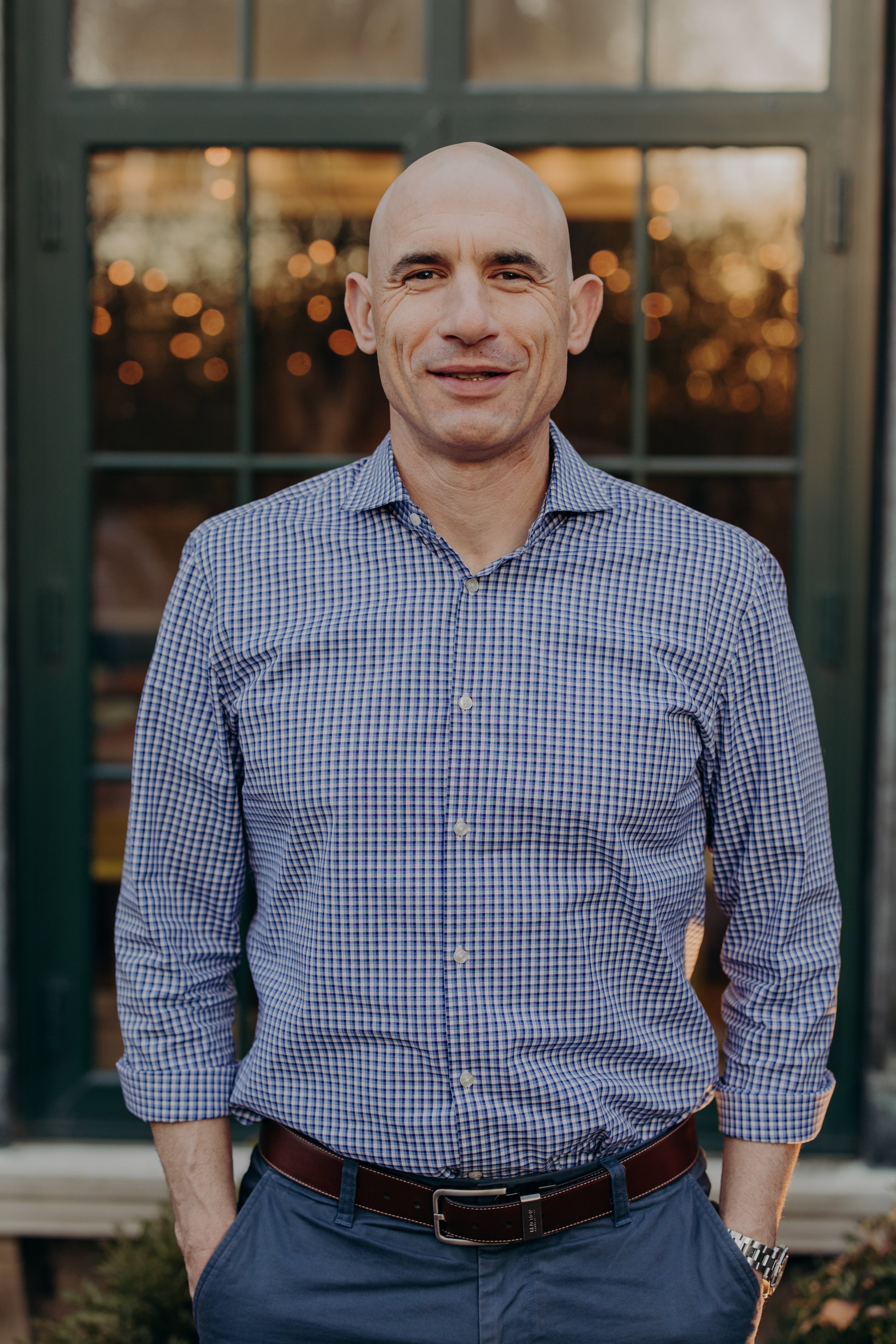 A man with a shaved head wearing a checked button-up shirt and blue pants, standing outdoors in front of a windowed building during sunset.