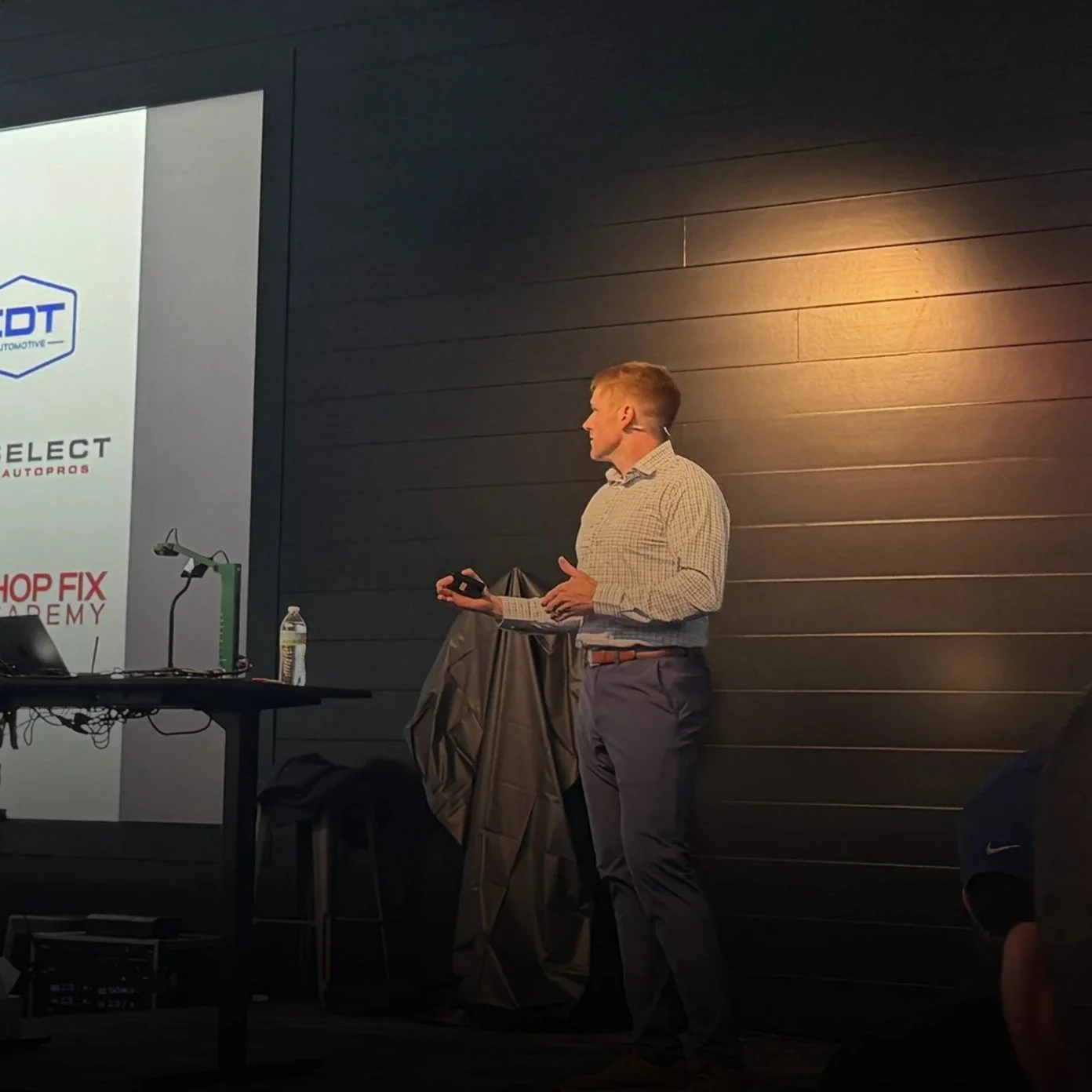 A man wearing a checkered shirt and gray pants giving a presentation in a dimly lit room with a dark wall. He is holding a remote control and has a microphone headset. A table with a water bottle, laptop, and equipment is nearby, and a large screen displaying logos is on the left.