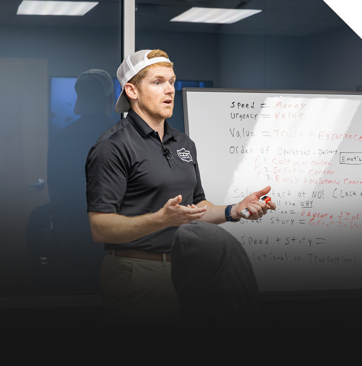 A man in a black polo shirt and white baseball cap giving a presentation in front of a whiteboard inside a conference room.