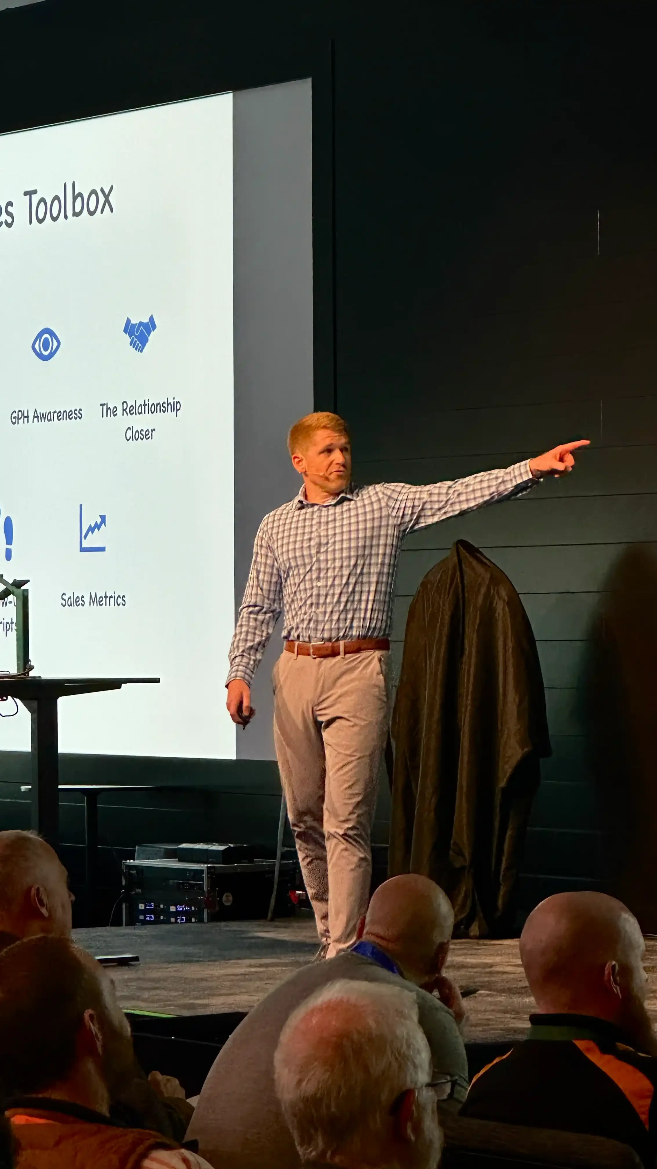 A man in a checkered shirt and beige pants giving a presentation on stage, pointing to a large screen displaying a slide titled 'Tools Toolbox' with icons and text related to GPH awareness, relationship closeness, sales metrics, and scripts; audience members are visible in the foreground.