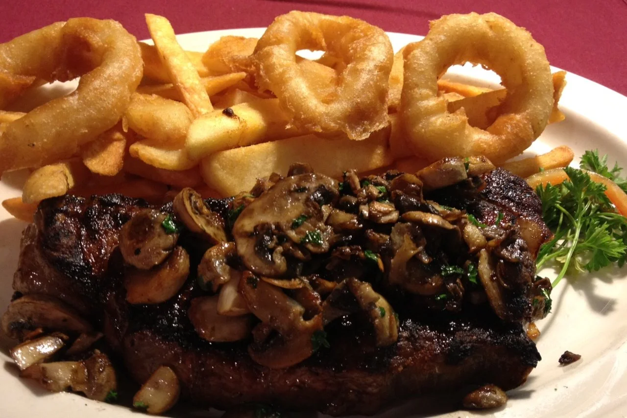 Steak topped with sautéed mushrooms and herbs, served with French fries, onion rings, and a sprig of parsley on a white plate.