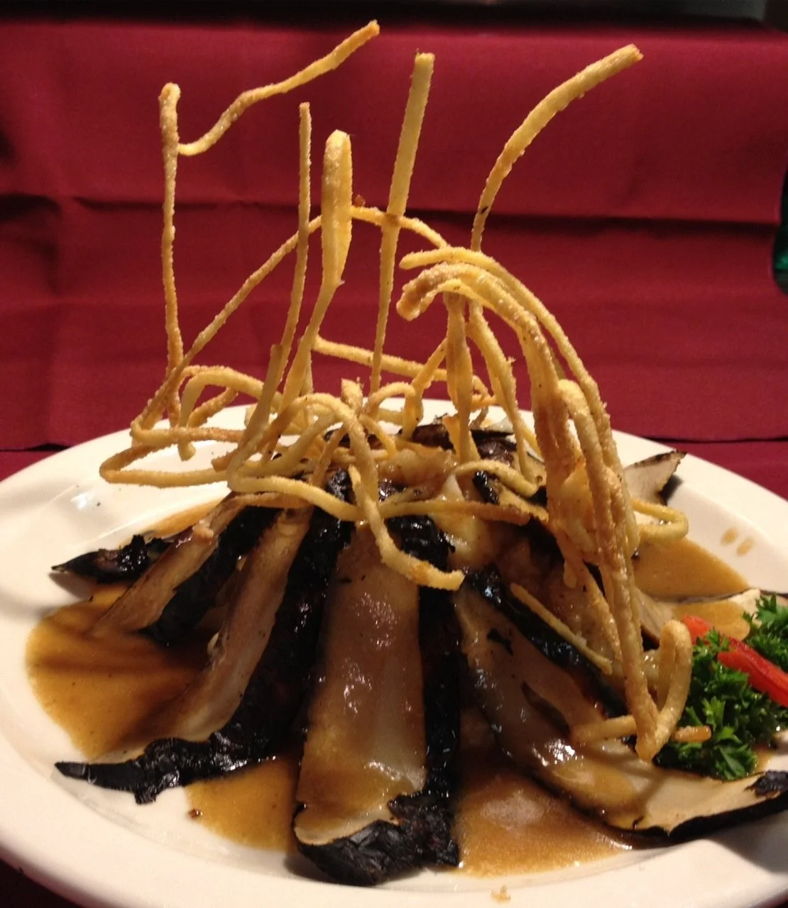 A plate of food with roasted eggplant slices, crispy fried noodles, and garnishes of parsley and red pepper, with sauce underneath.