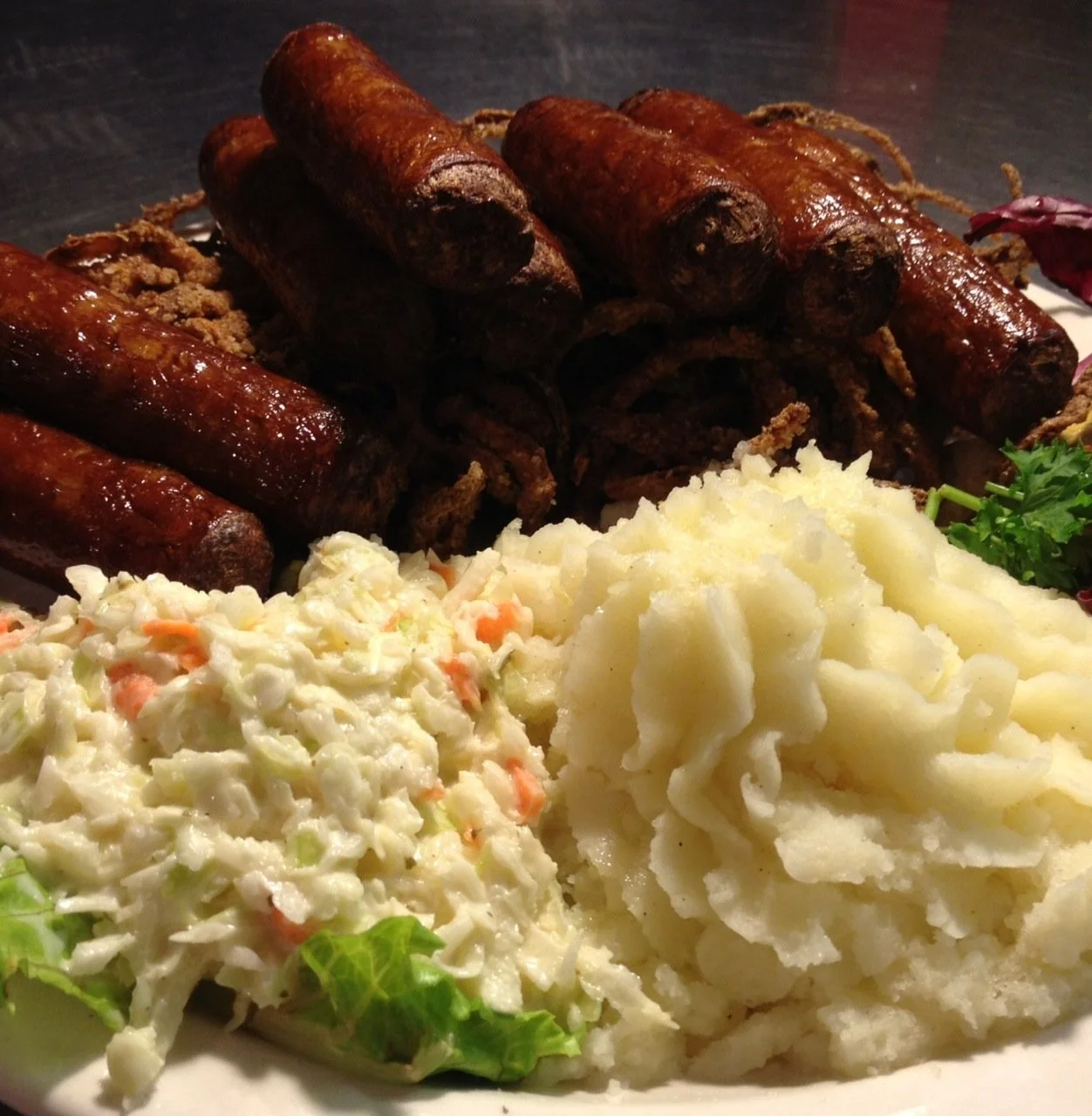 Plate of grilled sausages, mashed potatoes, and creamy coleslaw garnished with parsley.
