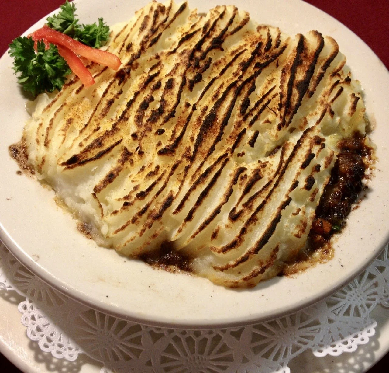 A plate of mashed potatoes with gravy, topped with toasted lines of what appears to be a dark sauce or gravy, garnished with parsley and a small piece of red pepper.