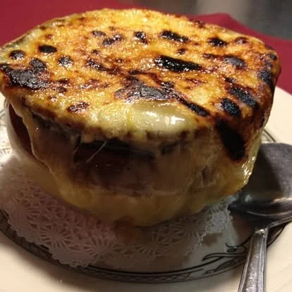 A bowl of French onion soup with melted cheese and toasted bread crust on top, served in a white bowl on a plate with a spoon.