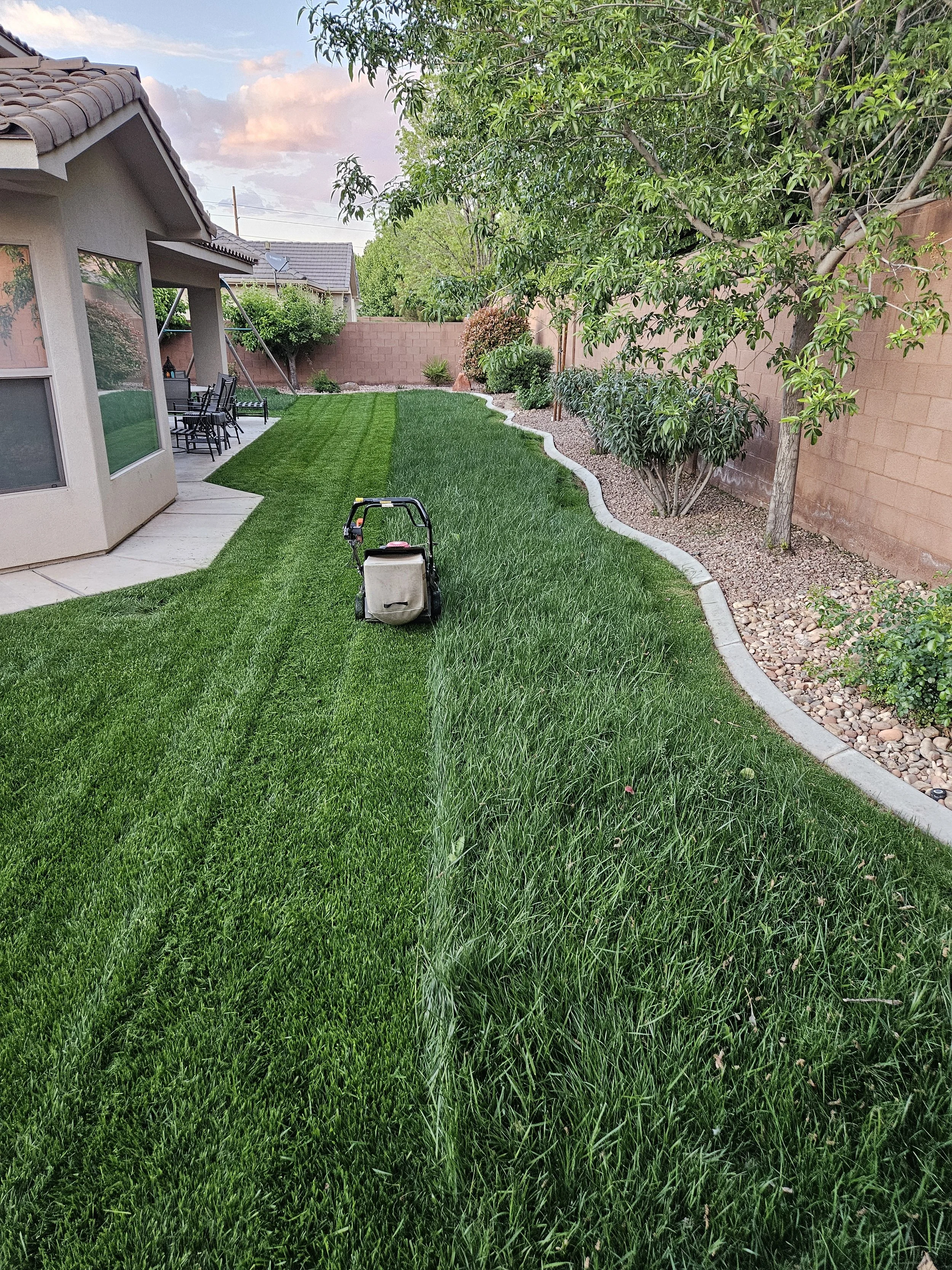 Backyard with green grass, trees, bushes, a brick wall, a patio with outdoor furniture, and a lawn mower.