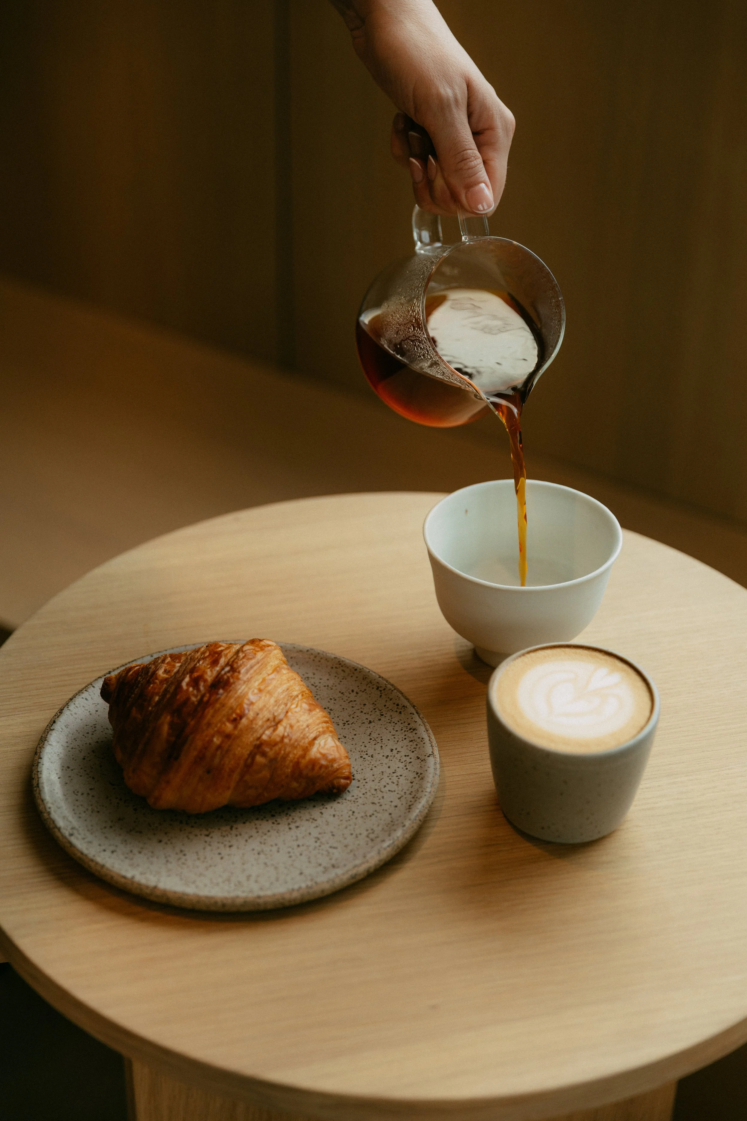 A hand pouring coffee from a glass carafe into a white cup, with a croissant on a speckled plate and a latte with foam art on a wooden table.