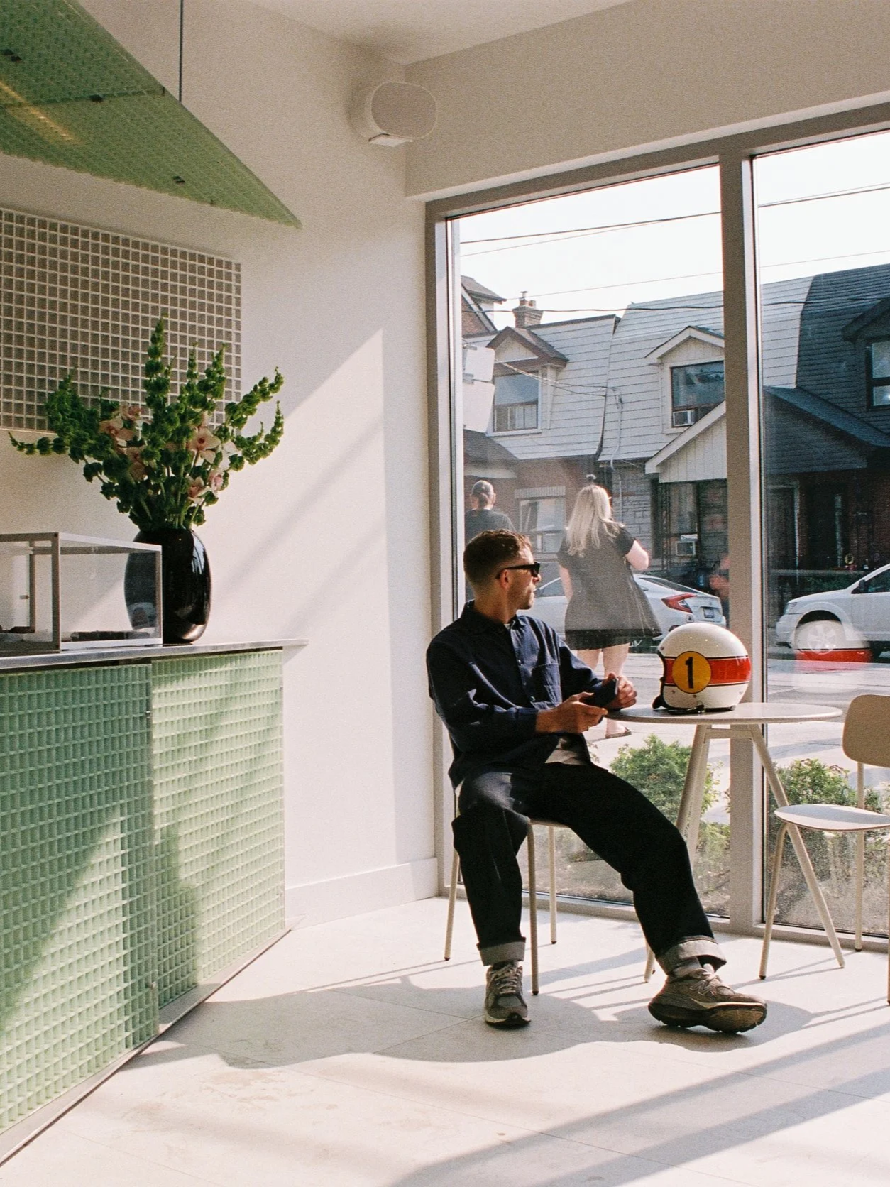 A man sitting inside a bright modern cafe near a large window, holding a smartphone and wearing sunglasses. On the small table in front of him is a white helmet with a racing number "1". Outside the window, people walk on the street and houses are visible.