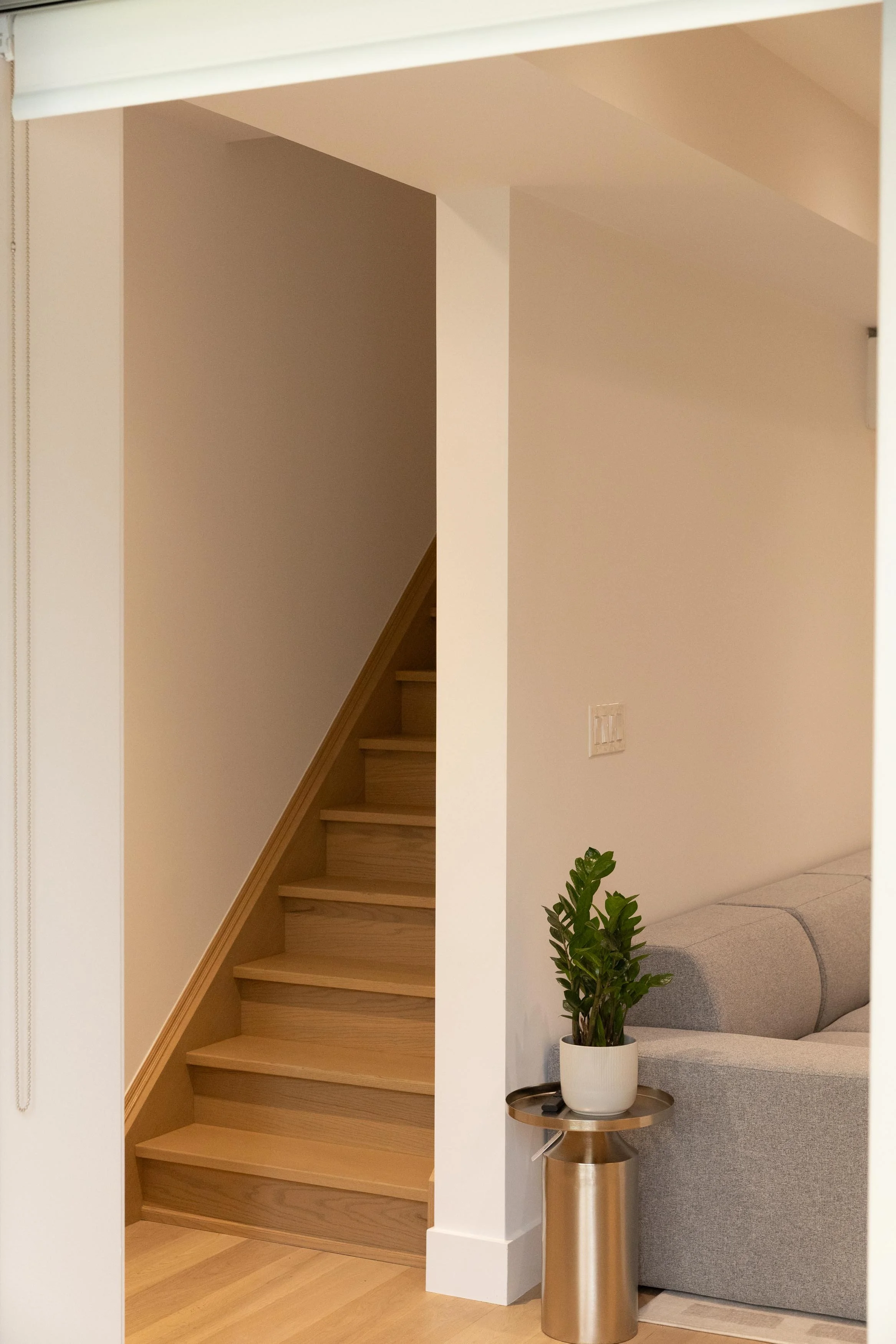 Wooden staircase inside a house with beige walls, next to a seating area with a gray couch, a potted green plant on a small table, and a wall-mounted light switch.