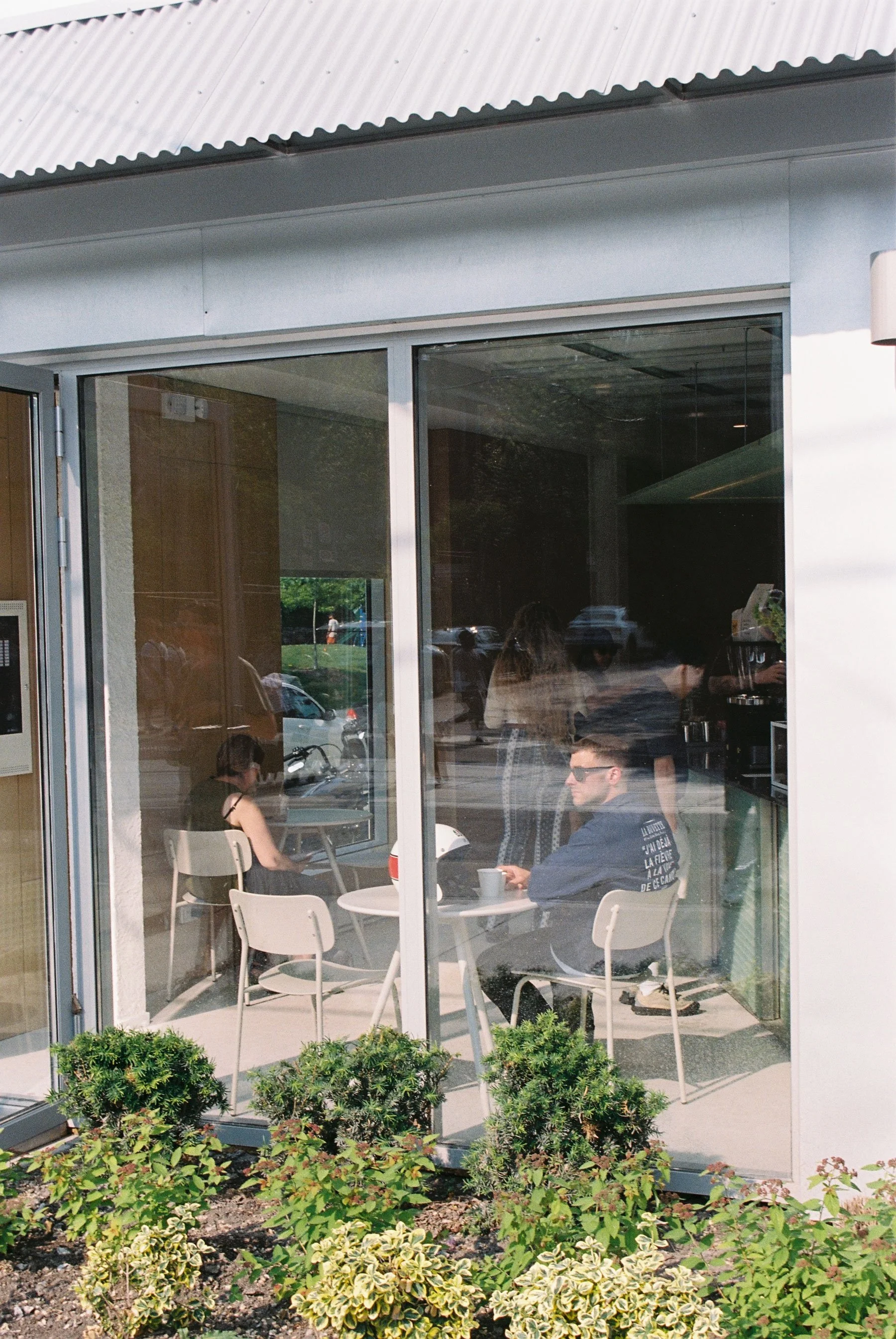 People sitting outside a cafe through a large glass window, with some greenery outside.